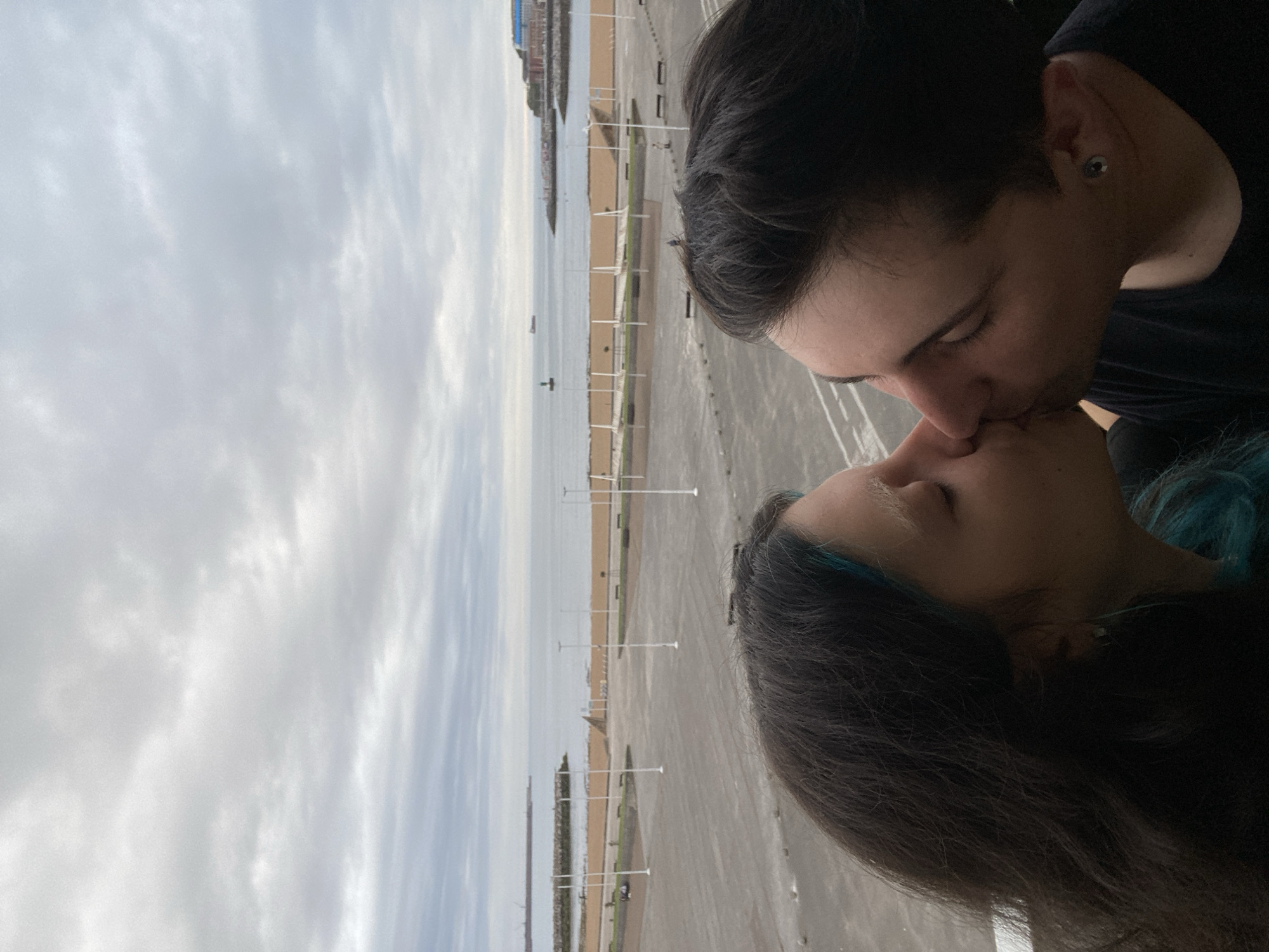 A photograph captures a couple in an embrace, their lips meeting in a kiss, set against the backdrop of Gijón/Xixón, Spain. The man, positioned on the upper right of the frame, has short, dark hair, with visible ear piercings. The woman, located at the bottom of the frame, has dark hair with streaks of blue. They are positioned in a way that suggests they are on an elevated platform, overlooking a beach.
In the background, the scene unfolds to reveal a stretch of beach bordered by the ocean and a paved area. Several white poles are spaced along the beach, likely serving as markers or supports. The sky overhead is overcast, with a predominance of grey clouds. The coastline extends in the distance, with buildings visible in the background. The paved area suggests the presence of a promenade or walkway, adding an urban element to the coastal setting.