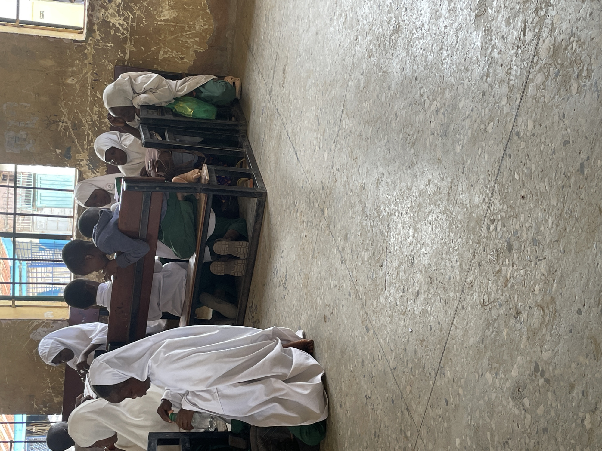 A classroom scene in Unguwar Rimi, Nigeria. Several students, partially visible, sit at desks arranged in rows. Some are wearing white gowns and head coverings. They appear to be focused on their work or reading. The room has tiled floors and distressed walls. Light comes in through windows on the left side.