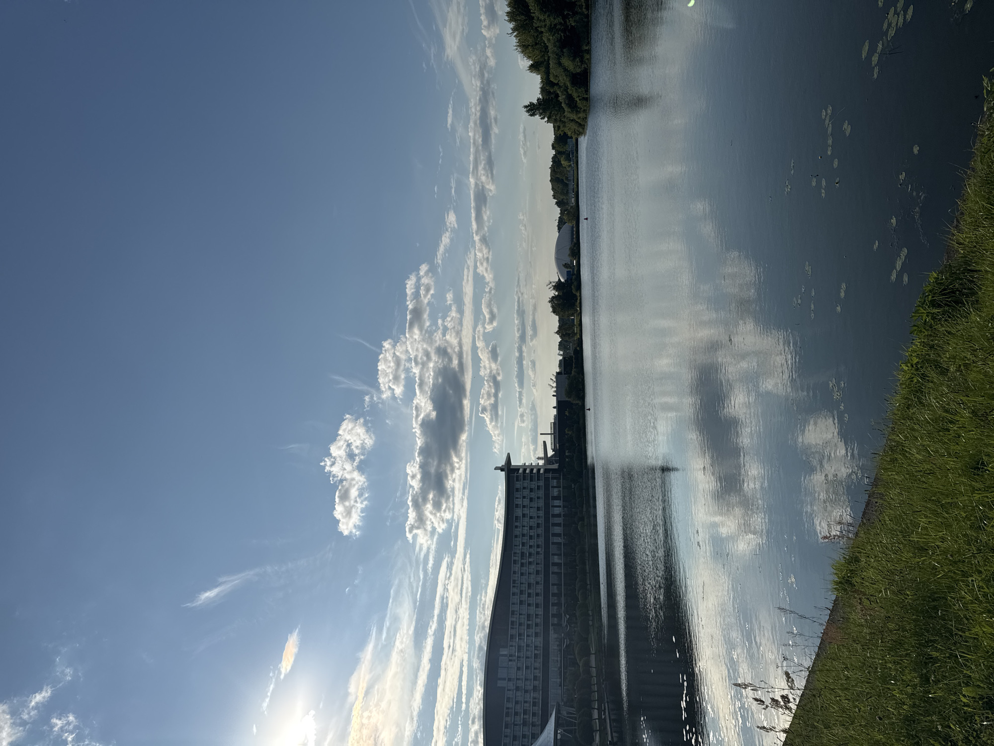 The image depicts a wide, calm body of water reflecting the sky and surrounding environment. On the left side of the frame, a large, modern hotel building with a curved facade and many windows stands along the water's edge. Trees line the area between the hotel and the water. The water itself is dark and smooth, with some lily pads visible on the surface in the foreground. The right side of the scene features a dense line of green trees. The sky is a clear blue with scattered white clouds, and the sun is visible on the far left, casting a bright light. The scene suggests a waterfront location, possibly a park or recreational area within a city.
Based on the provided information, this image is likely located in Minsk, Belarus. The hotel is identifiable as the "Minsk Marriott Hotel" or "Renaissance Minsk Hotel" which are situated along the Svislach River. The architectural style of the hotel and the presence of the river and green spaces are consistent with the Minsk waterfront.