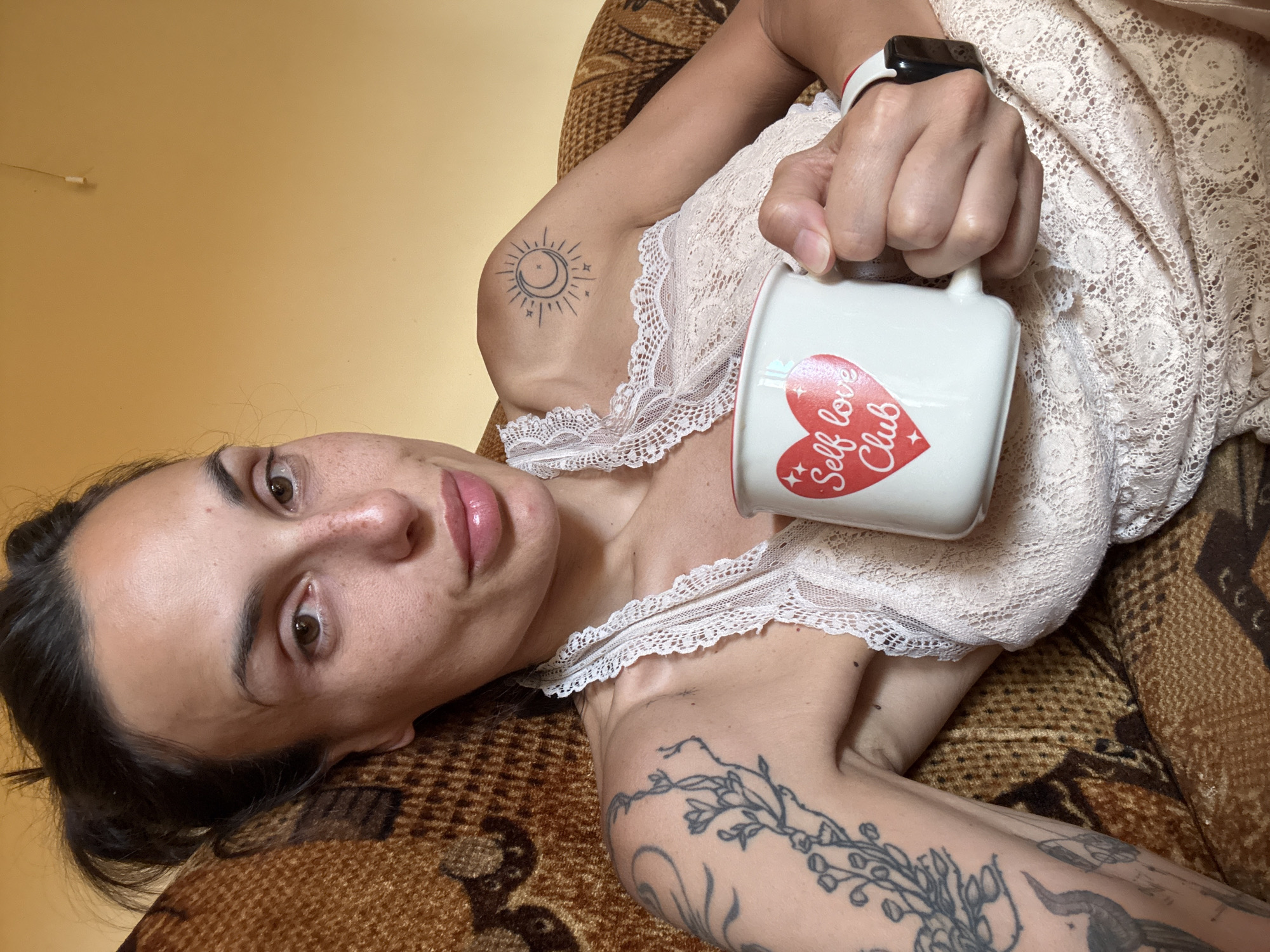 A woman with dark hair pulled back sits in a brown patterned armchair, holding a white mug with a red heart and text that reads "Self love Club." She is wearing a light-colored lace top. Tattoos are visible on her left arm and shoulder, including a floral design and a sun and moon symbol on her shoulder. Her right wrist has a black smartwatch. The background is a plain yellow wall. The scene appears to be an indoor setting in Pleven, Bulgaria.