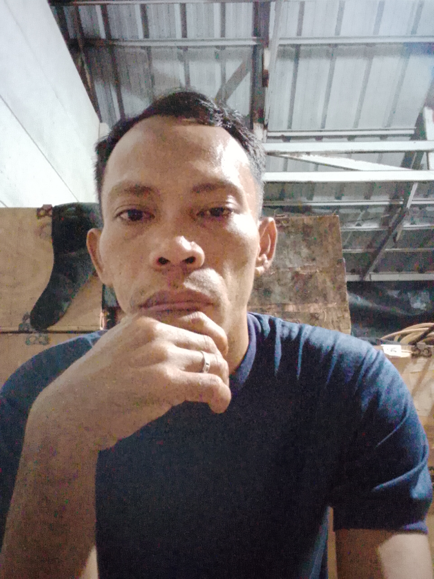 A man is pictured in a medium shot, indoors, with his hand resting on his chin. He is wearing a dark blue t-shirt. The background is out of focus, but appears to be an interior space with a corrugated metal ceiling and wooden structures. There is a ring on the man's finger. The overall lighting suggests it may be evening or a dimly lit environment. The location is identified as Bukit Ketok, Indonesia, though no specific landmarks are visible to confirm this.