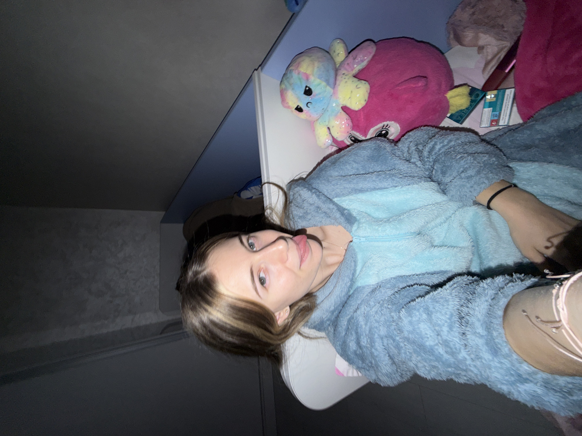 A young woman with light brown and blonde hair is lying in bed in a room. She is wearing a light blue, fluffy hooded jacket. Next to her on the bed is a pink plush animal with a multicolored octopus stuffed animal on top. Also on the bed are various other bedding items, including a pink blanket and a small rectangular item that could be a box or a book. The room appears dimly lit, with a gray wall visible in the background.
It is not possible to determine the location of the image in the city of Plovdiv, Bulgaria. The information is missing from the provided content.