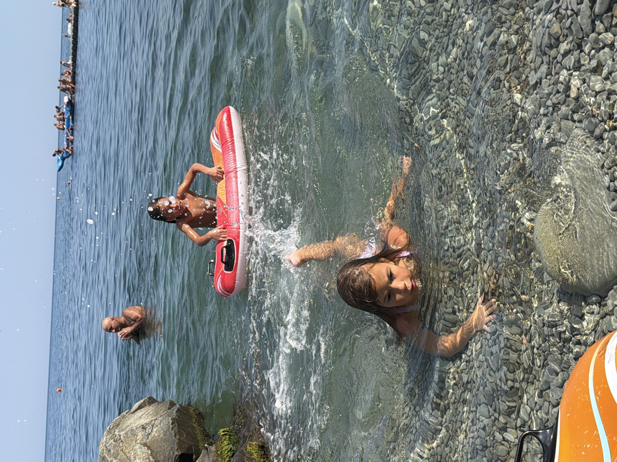 This image depicts a beach scene in Partenit, Ukraine. In the foreground, a young girl with long brown hair is swimming in shallow, clear water over a pebble-strewn seabed. She is partially submerged, with her head and arms visible. Further out in the water, another young girl is sitting in a red and white inflatable tube, her hands resting on its edge. In the background, several people are visible in the sea, some of whom appear to be on a long, inflatable watercraft. A bald man with his hands clasped is standing in the water to the left of the frame. The water is calm and blue, with a clear sky above. The overall environment suggests a sunny day at the beach.
