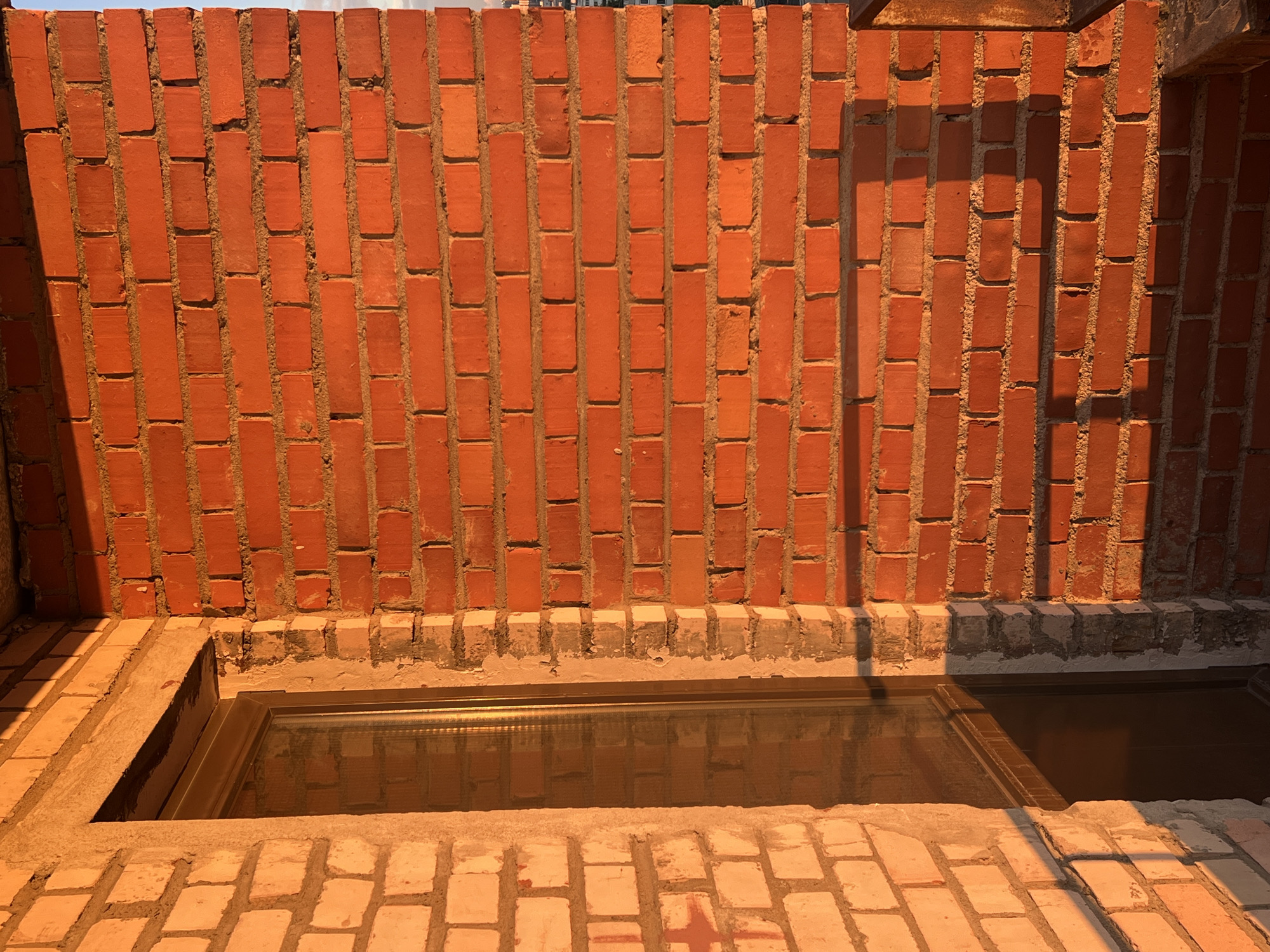 The image shows a close-up view of a brick wall and a skylight. The wall is constructed from reddish-orange bricks with visible mortar lines. The bricks are laid in a standard running bond pattern. Sunlight casts strong shadows across the wall, indicating it is either morning or late afternoon. The top of the image shows a sliver of blue sky, suggesting clear weather.
In the foreground, a rectangular skylight is set into a brick surface. The skylight has a metal frame and appears to be made of glass, reflecting the surrounding bricks and the brick wall. The area around the skylight is also paved with bricks, and there is a concrete structure surrounding the skylight opening. The overall impression is that of an outdoor setting, possibly a rooftop or patio area under construction or renovation. There is no visible text in the image. The scene is devoid of any people or discernible activity, focusing solely on the architectural elements and the play of light and shadow. The warm, golden light suggests a peaceful, quiet moment.