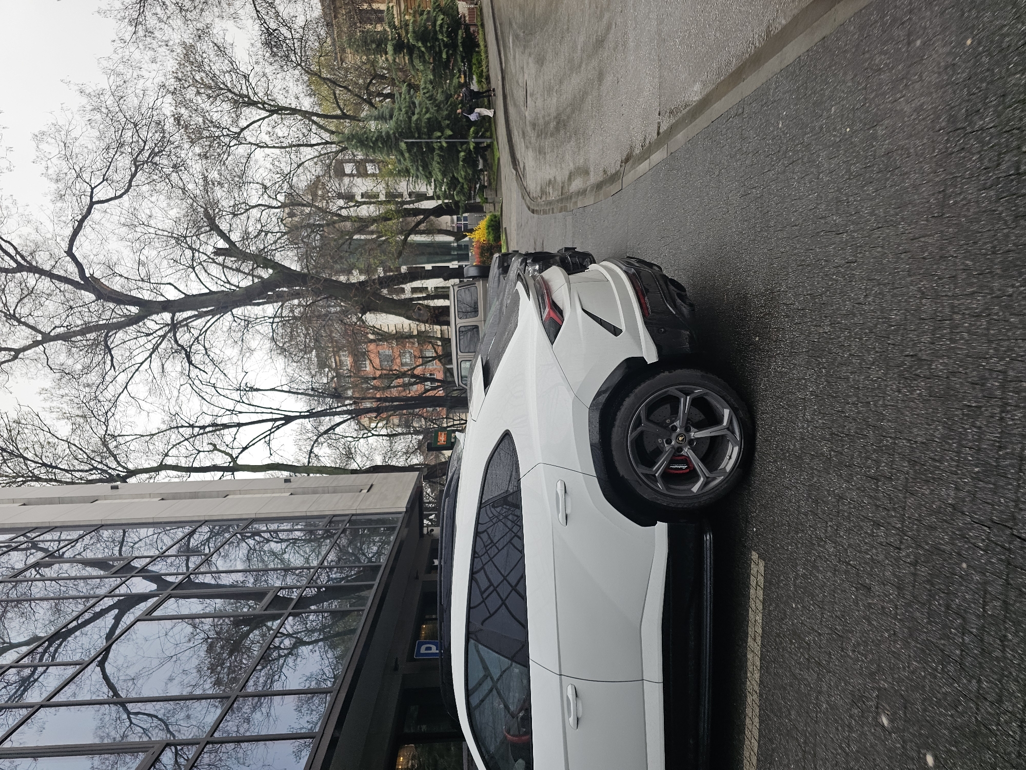 A white Lamborghini Urus is parked on a wet city street in Plovdiv, Bulgaria, adjacent to a modern building with reflective glass windows. A tree with bare branches is visible in the background, along with other buildings and greenery. Two figures are walking on the sidewalk in the distance. Other cars are parked further down the street. A parking sign is visible next to the building. The street is wet, likely from recent rain.