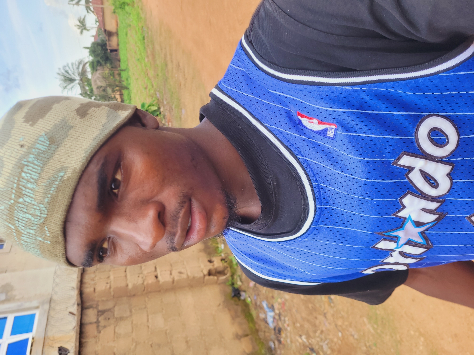 A young man, appearing to be of Nigerian descent, is pictured in a close-up, profile view, facing to the left. He is wearing a light green knit beanie with a camouflage-like pattern and a blue basketball jersey over a black t-shirt. The jersey has white trim and features the NBA logo and the word "Orlando" with a star. The background suggests an outdoor setting in a somewhat rural or developing urban area. To the left of the man is a light-colored building with a window featuring blue panes. Further back, there are more buildings, palm trees, and vegetation. The ground is a reddish-brown dirt path with some scattered debris. The sky above is partly cloudy. The image appears to have been taken in Bukuru, Nigeria, based on the style of the buildings and the surrounding environment.