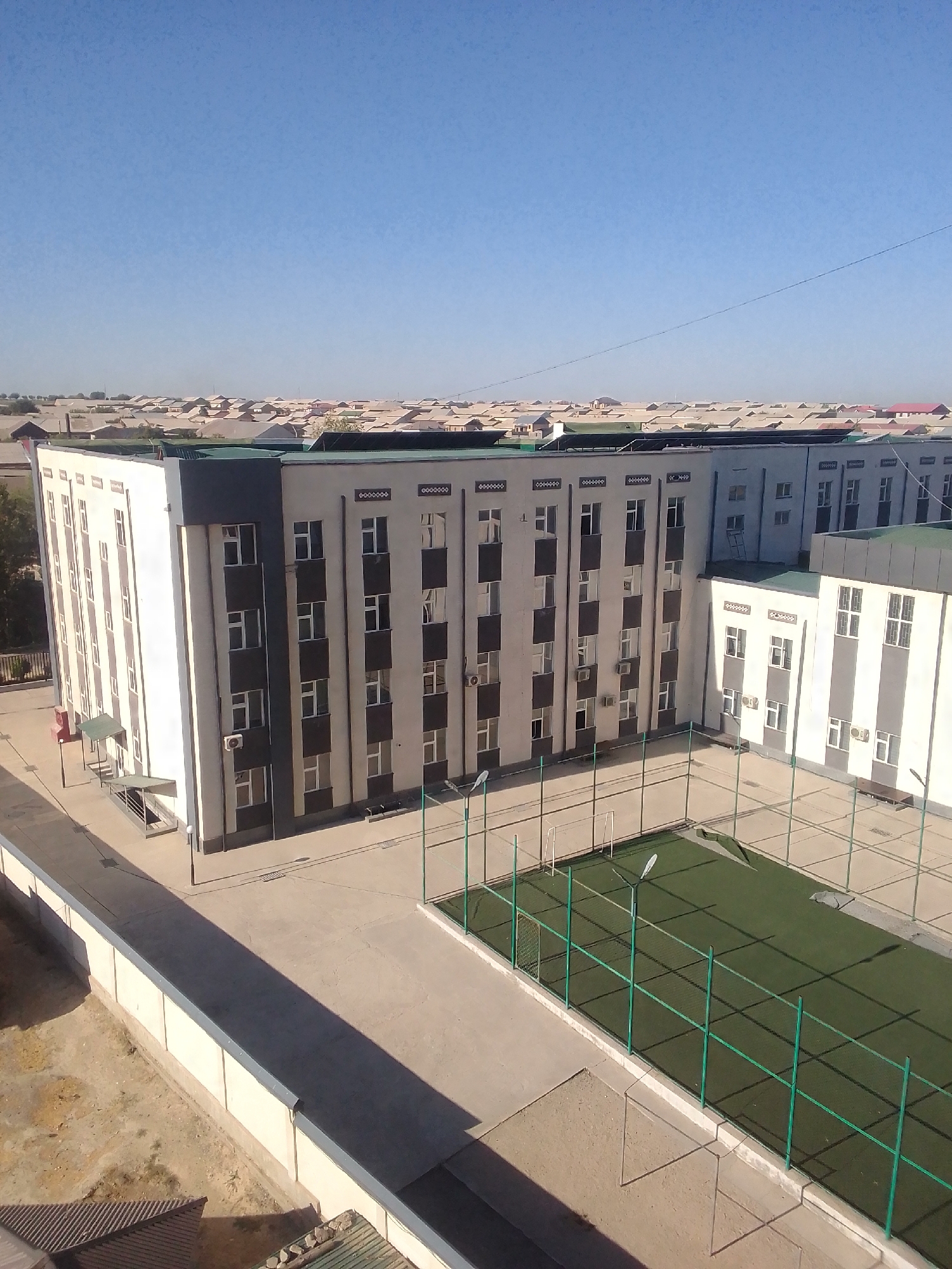 The image shows an aerial view of a modern building complex in Kattakurgan City, Uzbekistan. The main building is a multi-story structure with a light-colored facade and dark vertical accents. It has numerous windows and air conditioning units visible. Adjacent to this building is a sports field with artificial turf, enclosed by a green fence. In the background, a vast expanse of residential buildings with similar roof styles stretches towards the horizon under a clear blue sky. The overall environment suggests a well-developed urban or semi-urban area. There are no people visible in the image.