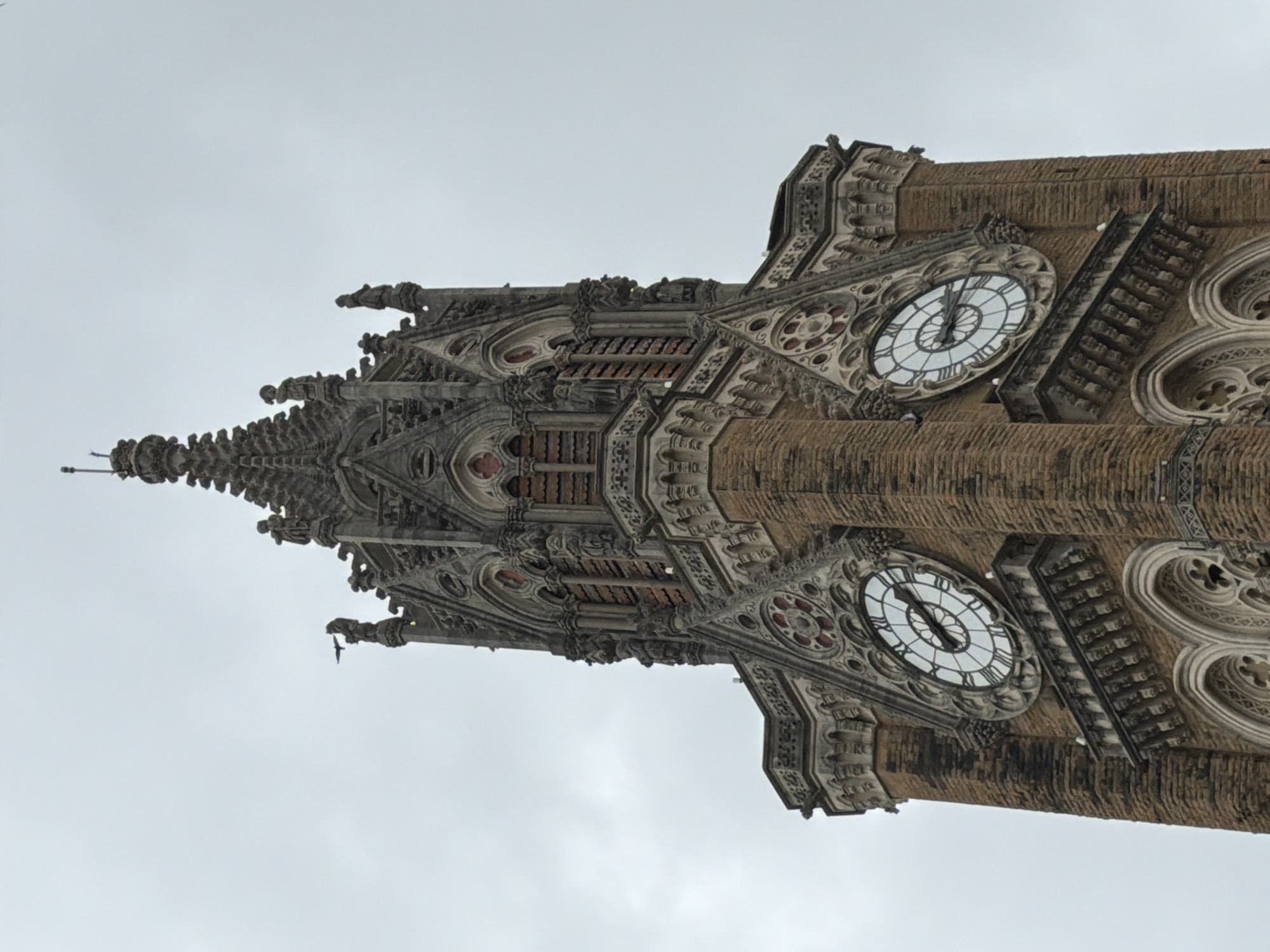The image is a low-angle shot of the Rajabai Clock Tower in Mumbai, India, under a cloudy sky. The tower, made of brown brick with ornate stone detailing, features a prominent clock face with Roman numerals. The clock shows the time as approximately 9:05. Statues of figures in traditional dress adorn the upper portions of the tower. A small bird is visible in flight near the top of the tower. The architectural style appears to be Gothic Revival.