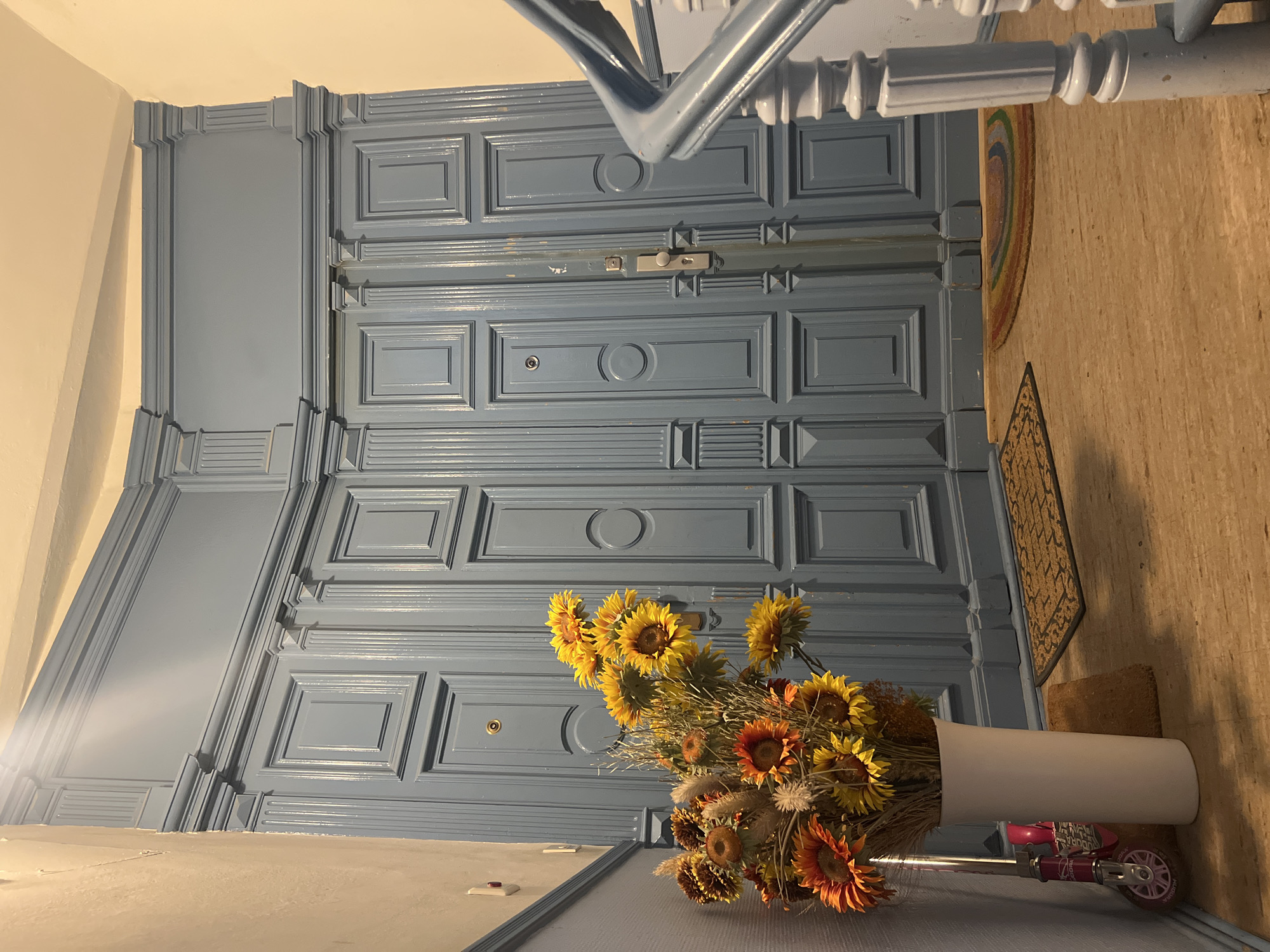 A hallway with ornate blue double doors in Berlin, Germany. The doors are intricately paneled with vertical fluting and decorative elements at the top and bottom. A silver doorknob and lock are visible on the right door. To the left of the doors, a large white vase holds a bouquet of sunflowers and dried flowers. Next to the vase, leaning against the wall, is a pink child's scooter. Two doormats are placed on the wooden floor in front of the doors. To the right, a light blue banister with white balusters is visible, part of a staircase. The walls are painted a light neutral color.