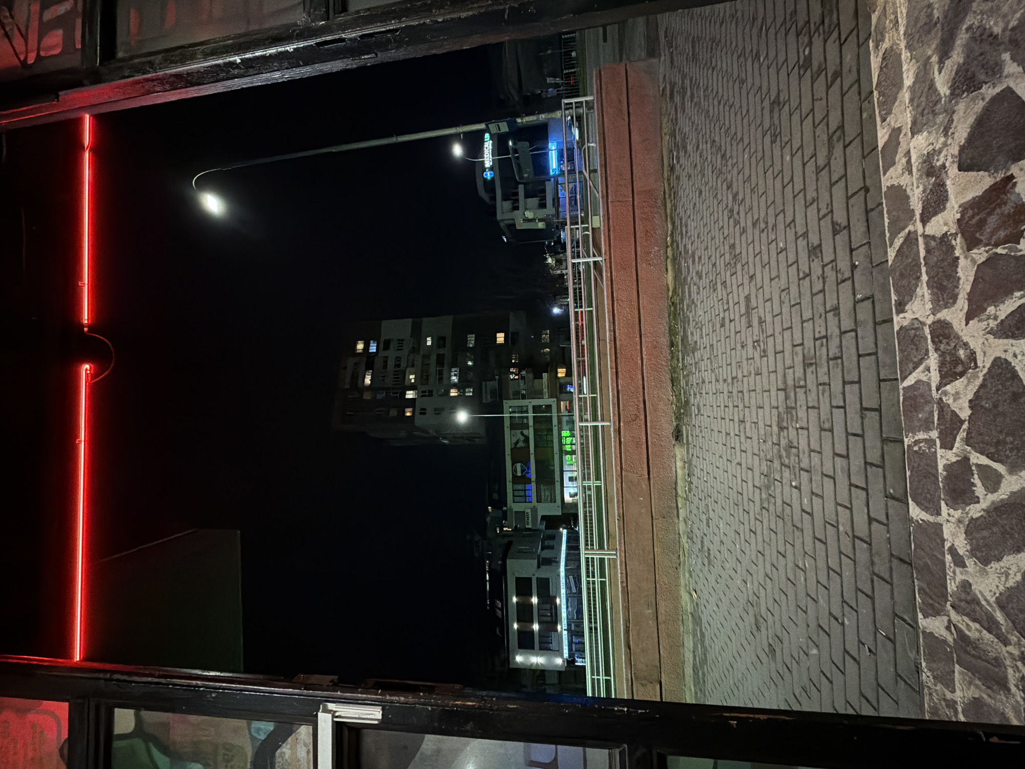 The image shows a nighttime view from an elevated position, likely a window or balcony, in a city that could be Varna, Bulgaria. A bright red neon light is visible to the left. In the distance, streetlights illuminate apartment buildings of varying heights. A street lamp in the foreground lights up a sidewalk paved with interlocking pavers, bordered by a stone wall to the right. The scene lacks people and direct interaction details, emphasizing urban architecture and the night environment.