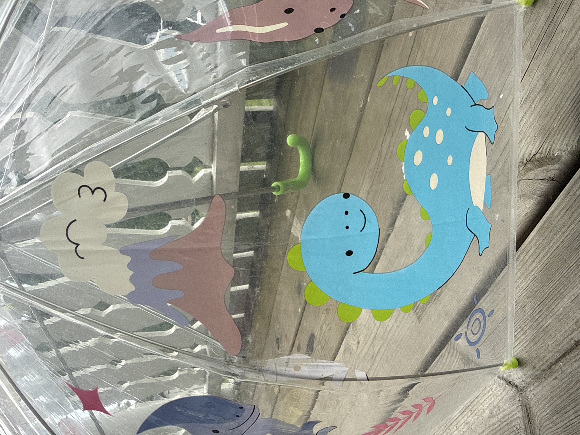 The image shows a close-up of a clear umbrella with cartoon dinosaur and volcano designs. The umbrella is positioned outdoors, resting on a wooden deck. In the background, there's a white railing and some greenery, suggesting a residential or outdoor public space. The umbrella features a blue dinosaur with green spikes and white spots, a pink starfish-like creature, a volcano with a smiling cloud and the number '3', and a red star. A small green plastic piece, possibly a handle or decorative element, is also visible. The overall impression is one of a child's accessory, likely used for rain protection. The image does not contain any information that would allow for its location to be specified in Ridabu, Norway.