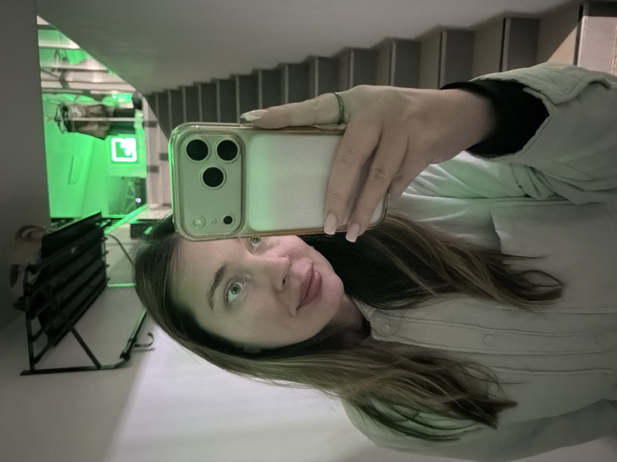 An adult female is depicted taking a selfie indoors, holding a gold-colored smartphone with a clear case in her right hand. Her gaze is directed towards the phone. She has long brown hair, light eyes, and a small nose ring in her left nostril. She is wearing a light-colored, possibly mint green or pale grey, collared garment with buttons and pleats on the front, and a dark lining visible at the cuff. A thin green ring is on her right ring finger, and her nails are manicured with light polish.
The background shows a light-colored interior space, likely a hallway or stairwell. To the upper left, a dark, wall-mounted shelf with two hooks is visible against a white wall. Directly behind and to the right of the subject, a light-colored staircase with darker treads ascends. The upper landing of the staircase is illuminated with a prominent green light, casting a strong green hue on the walls and a visible green neon sign, possibly displaying a stylized letter or symbol, on the far wall. The scene is an indoor environment. The specific location within the city of Chaikovsky, Russia, cannot be determined from the visual information provided in the image.
