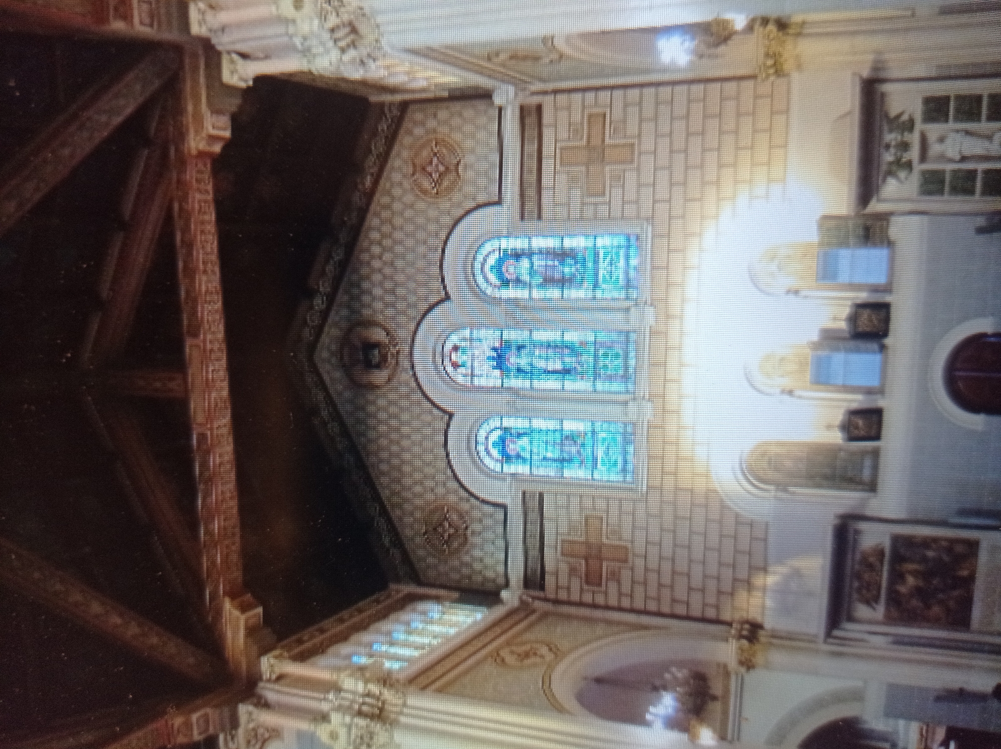The image shows the interior of a church, likely the Saint Joseph Church in Mukachevo, Ukraine, as indicated by its architectural style and stained glass windows. The focal point of the image is a series of three tall, arched stained glass windows with blue and yellow panes, depicting religious figures. These windows are set within a brick wall adorned with decorative patterns and cross motifs. Above the windows, the ceiling features exposed dark wooden beams, suggesting a historical construction.
To the left and right of the central section, ornate columns with decorative capitals rise, supporting the upper structure of the church. Below the stained glass windows, the wall transitions to a lighter color, with arched recesses and what appear to be smaller windows or decorative panels. In the foreground, on the left, a framed artwork or icon is visible. On the right, a tall, ornate structure with a statue and decorative elements is present. The overall impression is one of a well-maintained, traditional religious space.