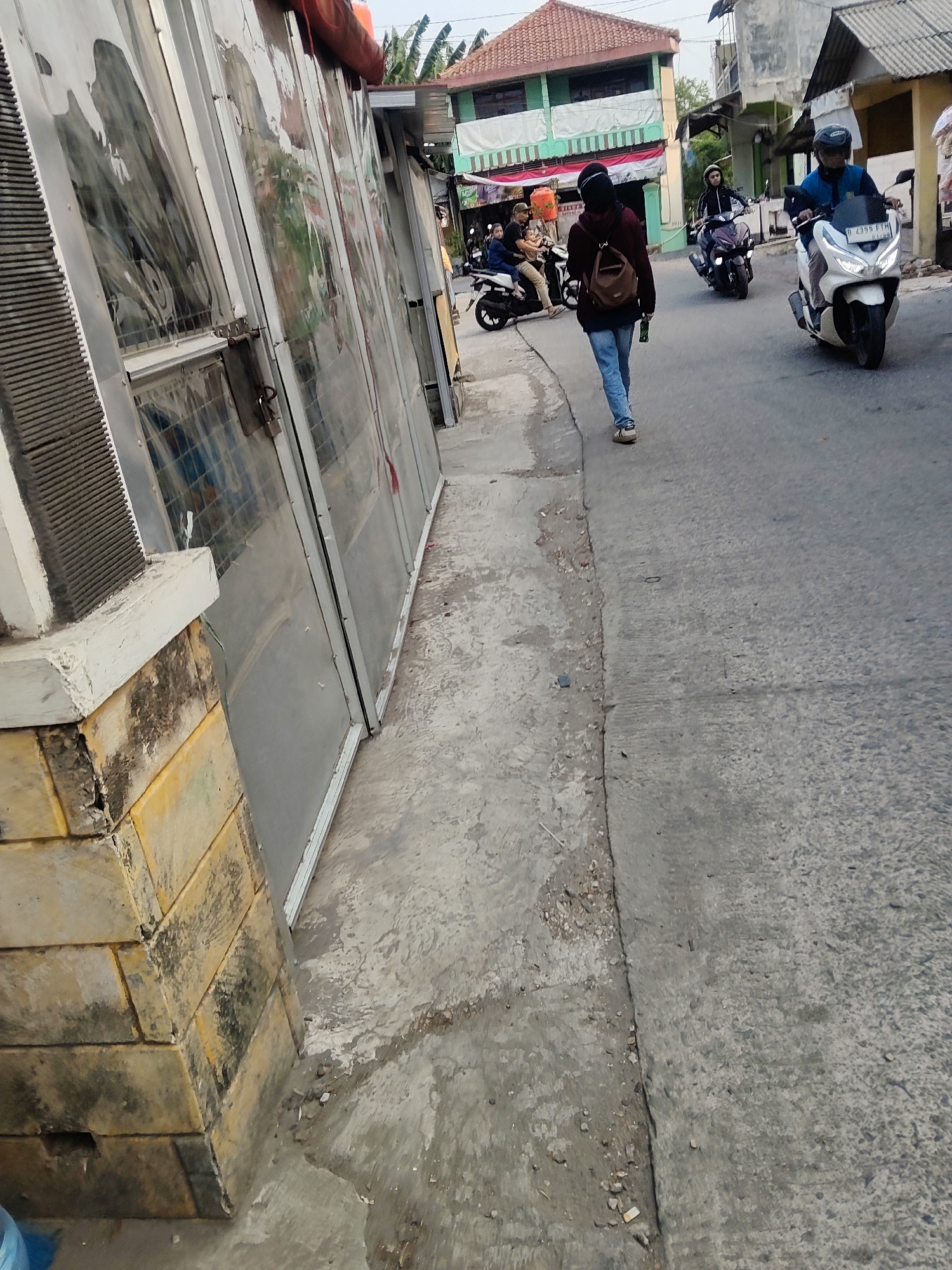 The image captures a street scene in Mekarmukti, Indonesia, with a focus on a woman walking on the right side of the road, heading away from the viewer. She is wearing a dark headscarf, a maroon jacket, and blue jeans, carrying a brown backpack. Several motorbikes are also present on the street. One motorbike with a rider in a blue jacket and helmet is approaching from the left. Another motorbike with a rider wearing a black jacket and helmet is further down the road to the left, also approaching. In the background, there are multi-story buildings, one with a green and white facade and a red tiled roof, and another lighter-colored building. The street is paved with asphalt, and there is a concrete sidewalk or pathway on the left side of the road, next to a structure with glass panels and a brick base. The environment appears to be a moderately urban setting, likely during daylight hours with the sun casting shadows.