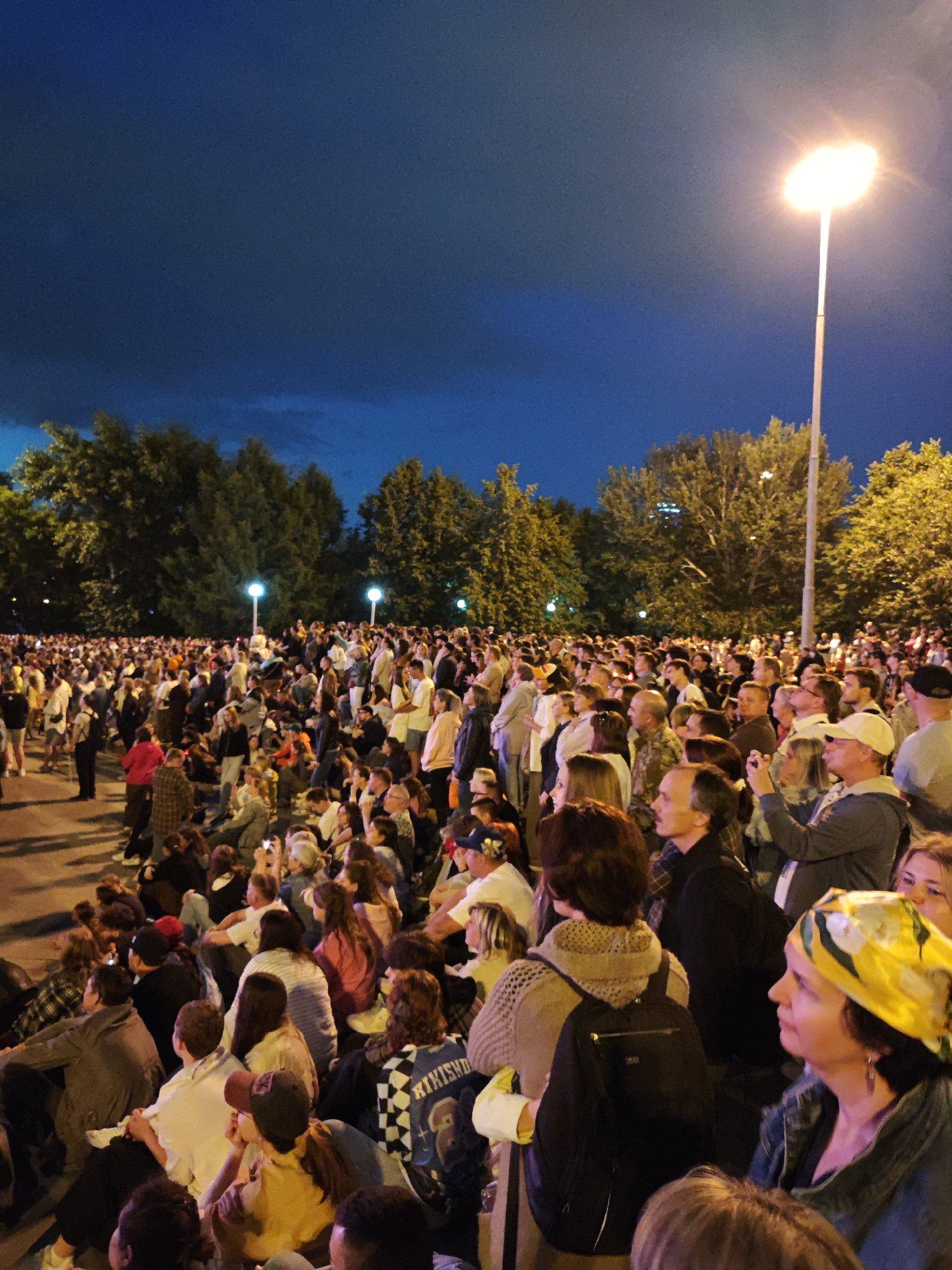 This is a wide-angle, low-angle outdoor photograph taken at dusk. A large crowd of people, estimated to be hundreds, are gathered in an open area, seated and standing. The sky is a deep blue, transitioning to black, with a large street lamp illuminating the right side of the frame with a bright, warm light. Trees with dense green foliage line the background. The people are diverse in age and are casually dressed. Many are looking in the same direction, suggesting they are watching an event or performance. Some individuals are holding up phones, possibly to take pictures or videos. The overall atmosphere appears to be one of shared experience and anticipation. The scene is likely a public gathering or festival.
