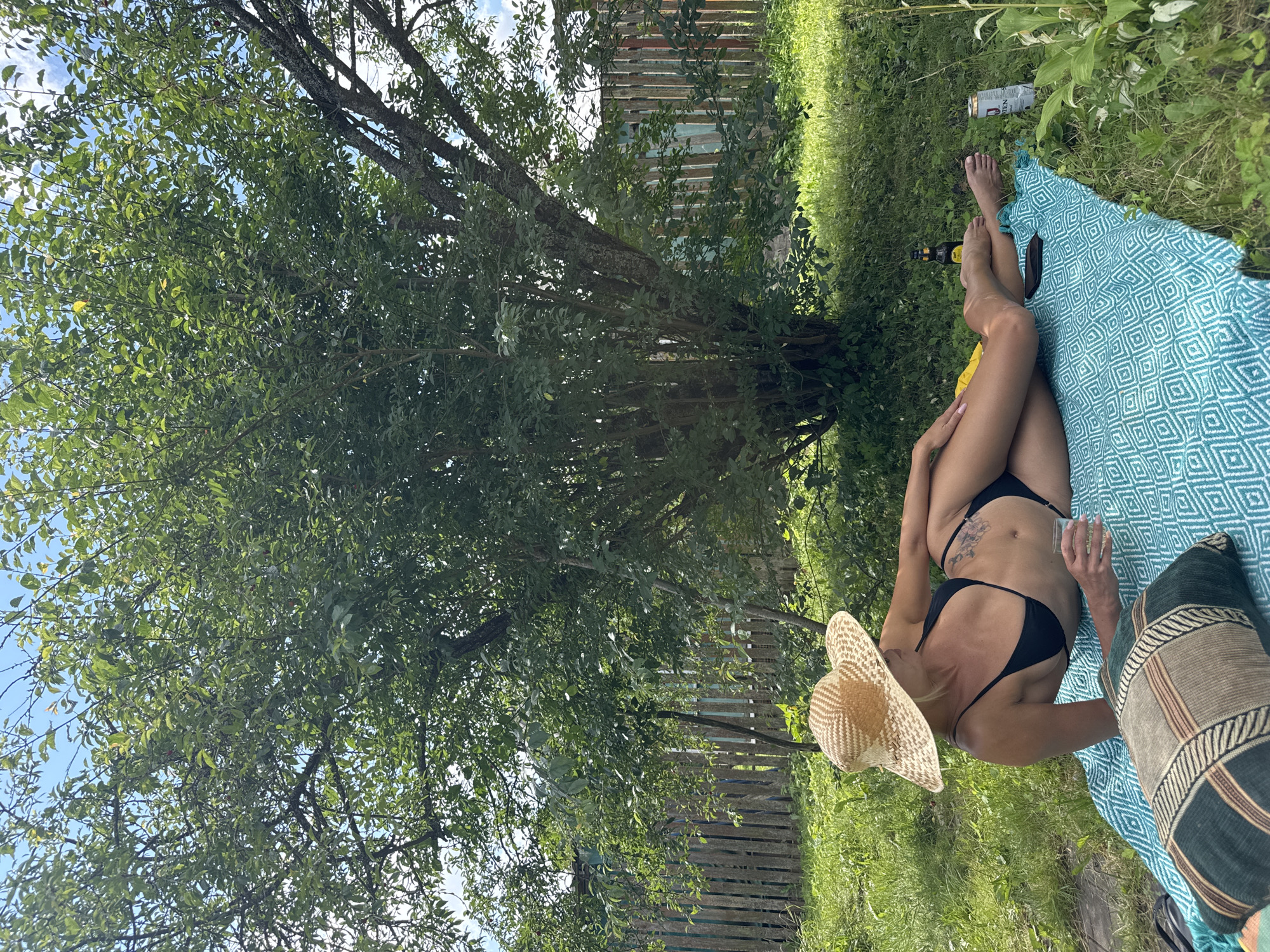 A woman in a black bikini and a straw hat lies on a patterned blanket on the grass under a tree. She is holding a glass of clear liquid in her right hand. To her right, on the blanket, are two bottles and a can. In the background is a wooden fence. The scene appears to be an outdoor setting, possibly a garden or backyard, in the city of Krasnaya Presnya SNT, Russia.
