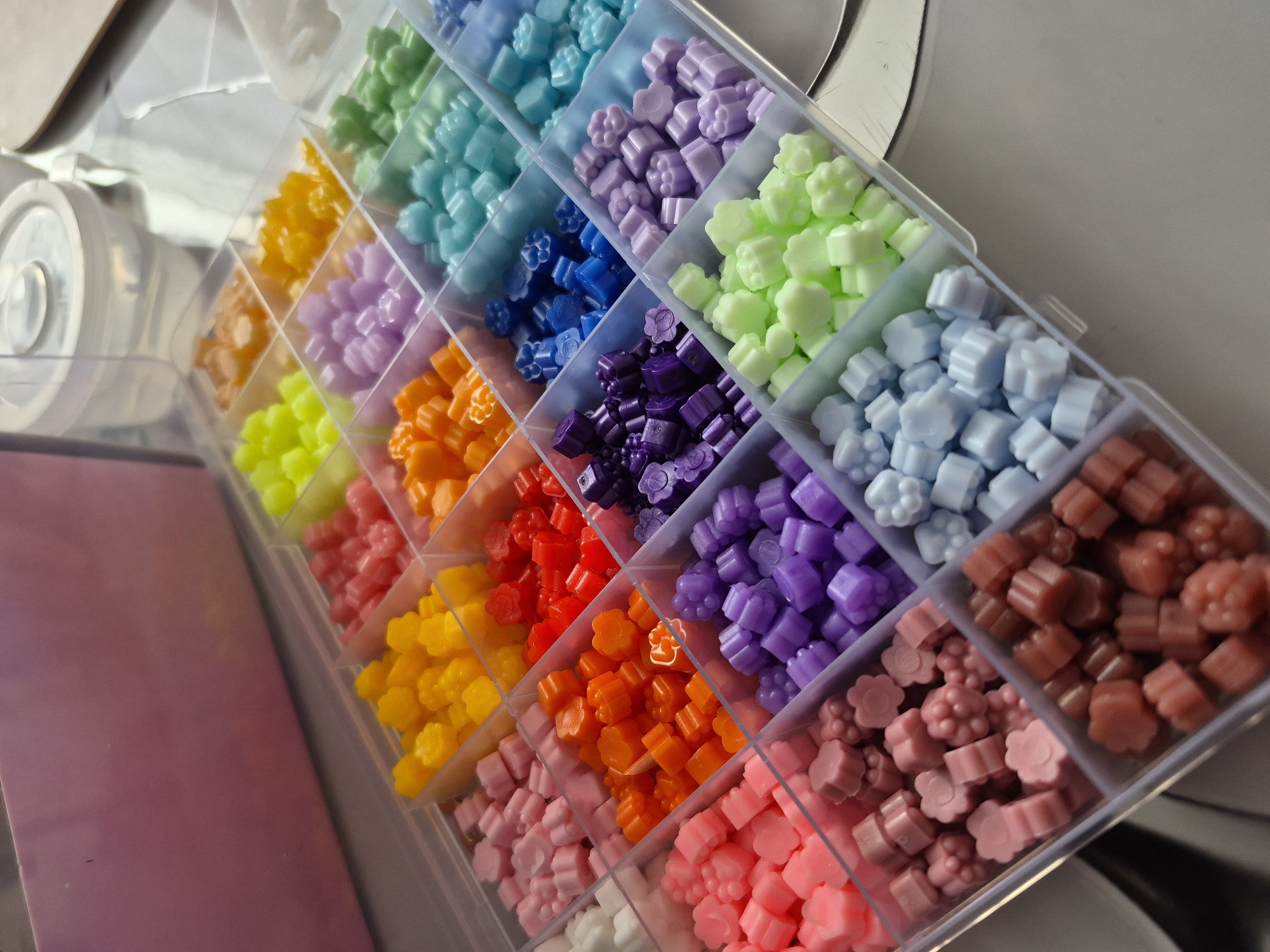 The image shows a plastic container with multiple compartments, each filled with small, colorful flower-shaped objects. The objects are arranged in a rainbow-like sequence of colors, ranging from light blue to green, yellow, orange, red, purple, and pink. The container is slightly tilted and sits on a surface that appears to be a light-colored tabletop or countertop. The background is slightly blurred, suggesting a shallow depth of field. The location is not discernible from the image. There is no evidence to suggest a location in Tobolsk, Russia.