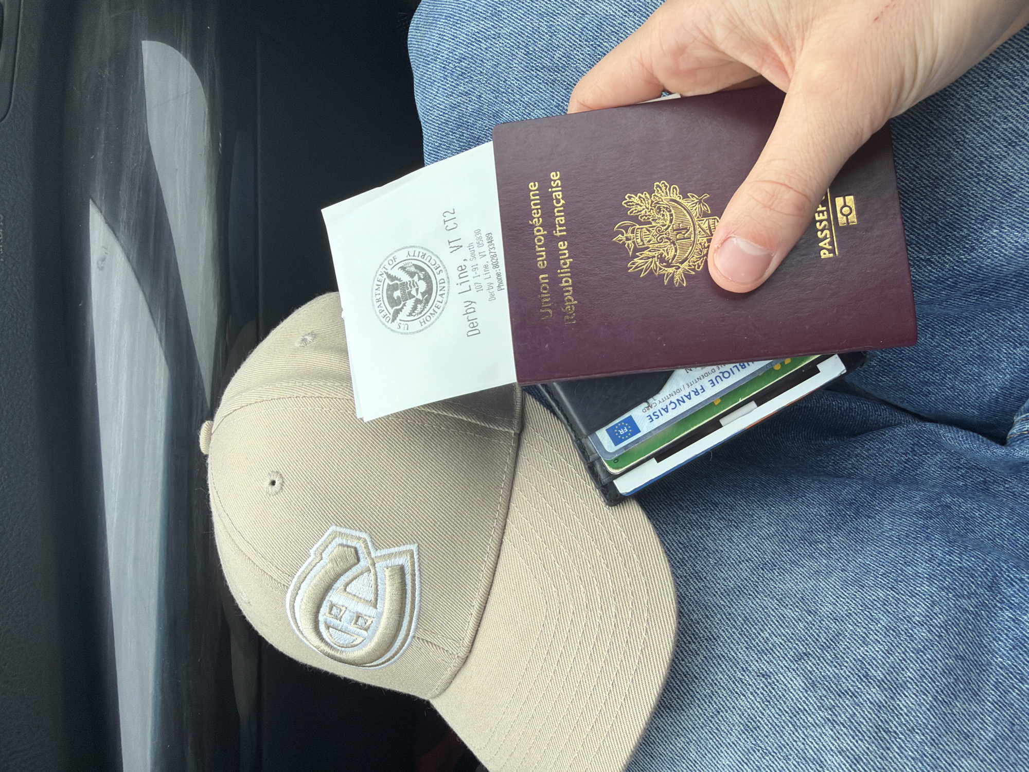Here is a description of the image:
A hand holds a French passport, a slim wallet with French identity cards, and a U.S. Department of Homeland Security document on top of a tan baseball cap resting on blue jeans. The Homeland Security document is from Derby Line, Vermont (VT CT2, 107 I-91 South, Derby Line, VT 05830). The background includes a blurry gray interior.
