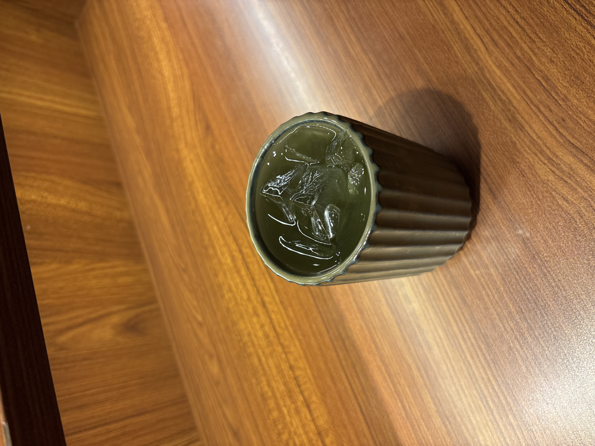 A dark green, ribbed ceramic cup filled with a clear liquid and ice cubes sits on a polished wooden surface. The background is a wooden shelf. The image was taken at a cafe in West Jakarta, Indonesia.