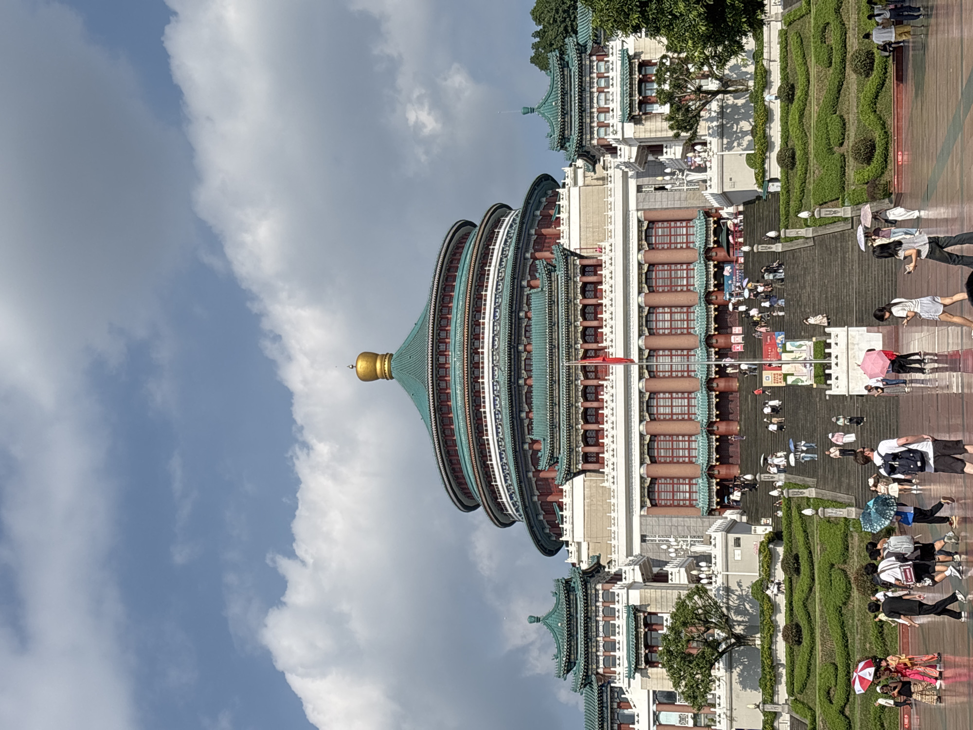The image shows the Chongqing People's Auditorium, a prominent landmark located in the Yuzhong District of Chongqing, China. The grand building features a distinctive multi-tiered, circular main hall topped with a green, domed roof and a golden finial. Flanking the main structure are lower buildings with similar traditional Chinese architectural elements, including layered green roofs and red columns.
In front of the auditorium, a wide, paved plaza reflects the cloudy sky. A large set of stairs leads up to the building, with numerous people scattered throughout the scene. Some individuals are walking across the plaza, while others are ascending or descending the stairs. Many people are carrying umbrellas, suggesting it might be sunny or there's a possibility of rain. The foreground shows a diverse group of individuals in casual attire, some walking in pairs or small groups, while others are alone. Landscaped green terraces with manicured bushes are visible on either side of the main stairway, adding to the formal aesthetic of the surroundings. The overall impression is of a busy public space on a partly cloudy day, with the impressive architecture of the People's Auditorium serving as the central focus.