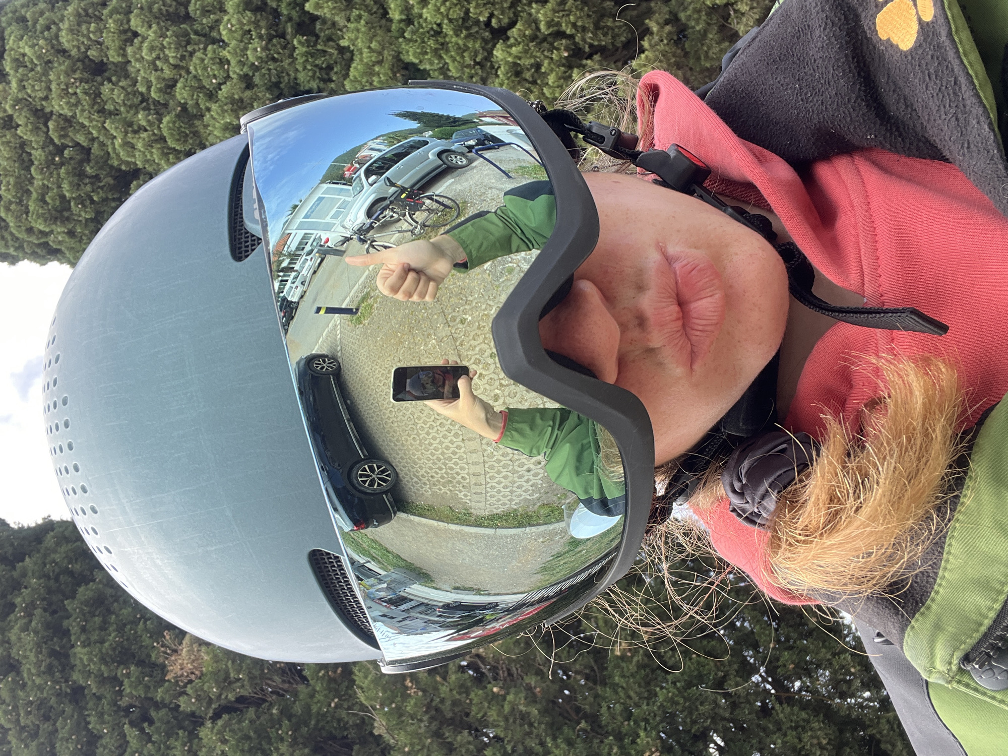 A person is wearing a helmet and goggles, taking a selfie. The helmet has a reflective visor, reflecting a thumbs up being given by the person in the image to a person using a phone in the reflection. Behind the person are parked cars and a bicycle on a paved surface. The scene is in Donji Pobori, Montenegro.