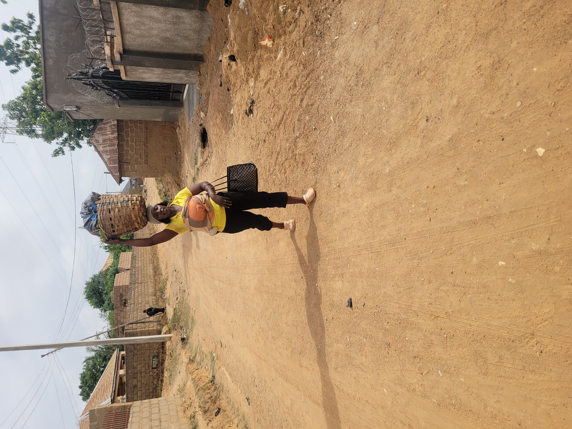 A woman is walking on a dirt road in Barakin Akawo, Nigeria. She is wearing a yellow shirt, black pants, and tan sandals. She has a basket on her head and is carrying a black plastic chair or other object. The road is unpaved and appears sandy. There are buildings visible on both sides of the road. Trash and vegetation are present on the side of the road. Overhead electric lines are visible.