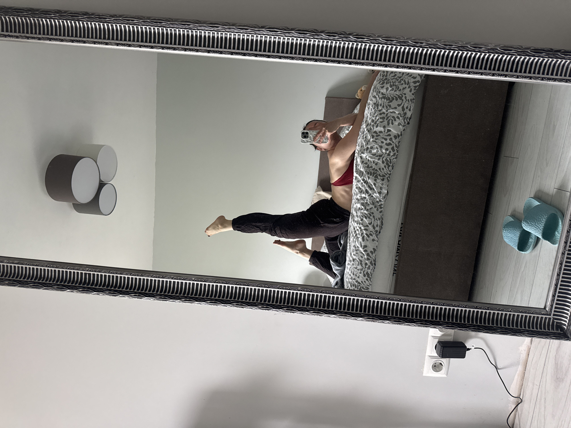 The image is a mirror selfie taken indoors, likely in a bedroom. A person, wearing a red bra and dark trousers, is reflected in a large, ornate mirror with a decorative silver and black frame. The person is lying on a bed, covered with a grey and white patterned duvet, and is holding a smartphone to capture her reflection. She is striking a pose with her right leg extended high upwards and her left leg bent.
To the left of the reflected person, three cylindrical wall-mounted lights are visible; two are dark grey/black, and one is white. The wall behind these lights is a light grey. The floor below the bed is light-colored wood. In the bottom right corner of the mirror's reflection, a European-style wall outlet is visible, with a black power adapter and cable plugged into it. Two light blue slide sandals rest on the floor next to the bed. The text "MAGIVA" is partially visible on the side of the bed frame in the reflection. The ambient lighting suggests it is daytime.