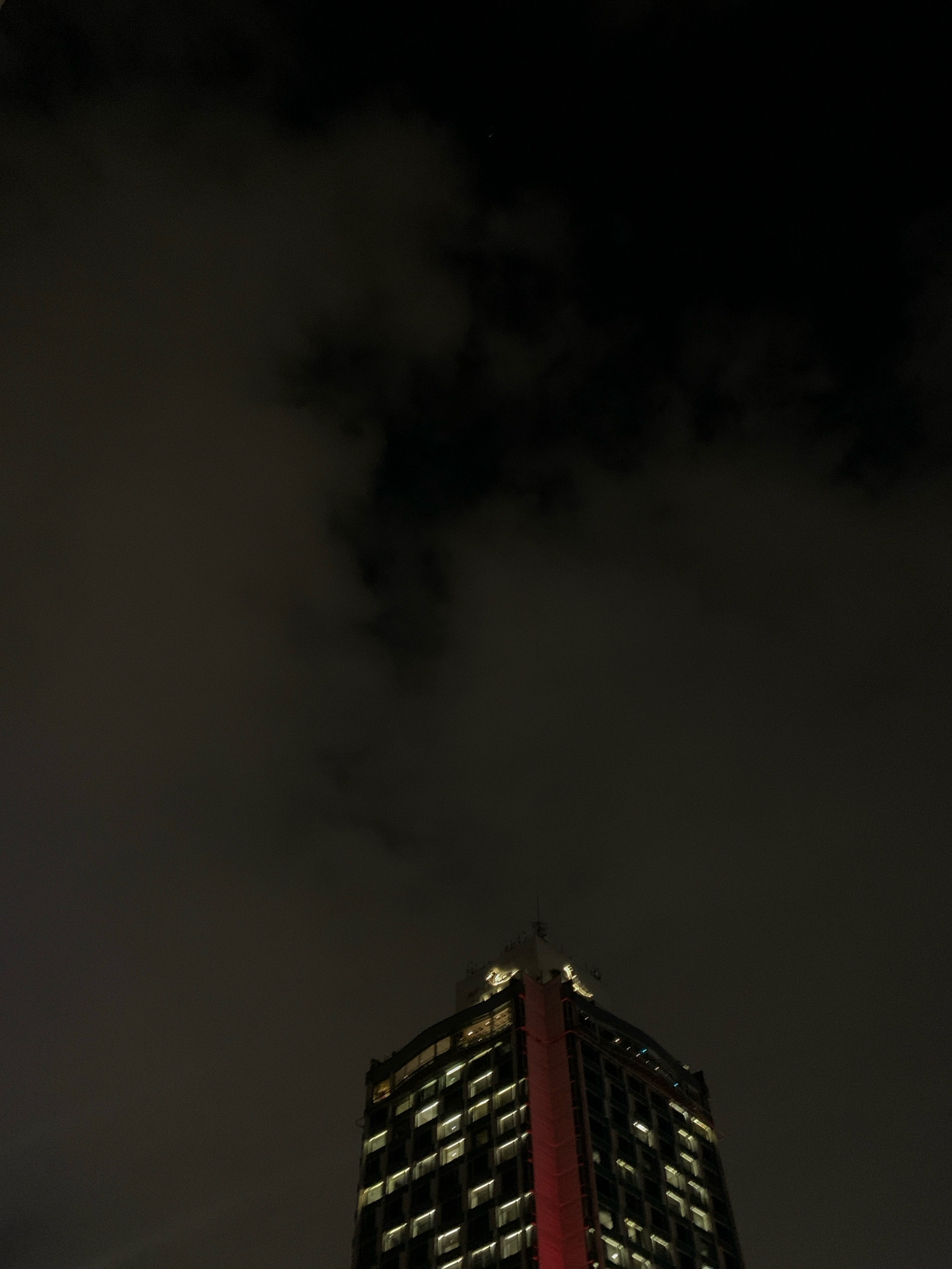 A low-angle, outdoor, nighttime photograph captures the upper portion of a modern, illuminated skyscraper in Beyoğlu, Turkey. The building is tall and features a grid of evenly spaced windows, many of which are lit from within, casting a warm glow. A striking vertical band of red light runs down the side of the building. The skyscraper's roof is visible, along with some antenna-like structures. The sky above is dark and filled with amorphous, dark gray clouds, obscuring any stars or moon. The overall atmosphere is somber and urban.