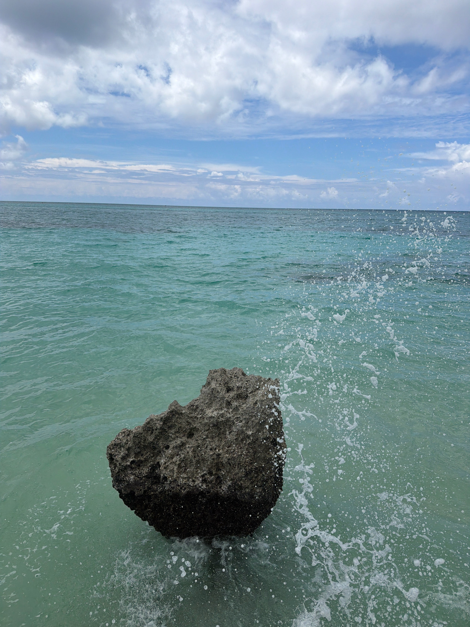 A rock is submerged in the turquoise waters of Malay, Philippines, with waves crashing against it and creating a spray of white foam. The sky above is a mix of blue with white clouds. The scene suggests a natural coastal environment.