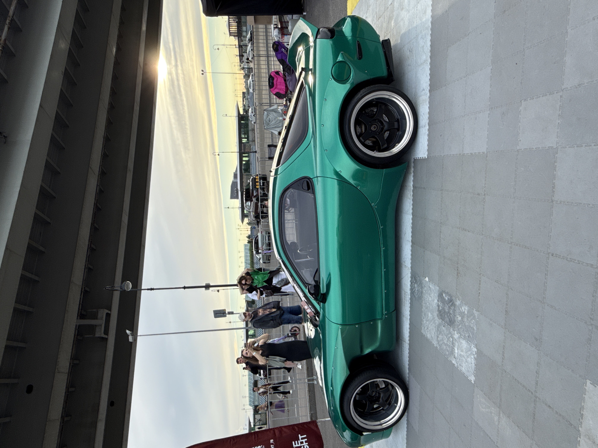 A modified, bright green sports car, possibly a Mazda RX-7, is displayed on a tiled surface outdoors. The car has wide fenders and a prominent rear spoiler. Several people are visible in the background, some taking photos of the car. A tall, dark banner stands to the left of the car. In the distance, buildings and a bridge are visible against a cloudy sky with the sun setting or rising, casting a warm glow. The scene appears to be an outdoor car show or event, possibly in Saint Petersburg, Russia, given the presence of a traffic light consistent with Russian street furniture and the architectural style of the distant buildings.