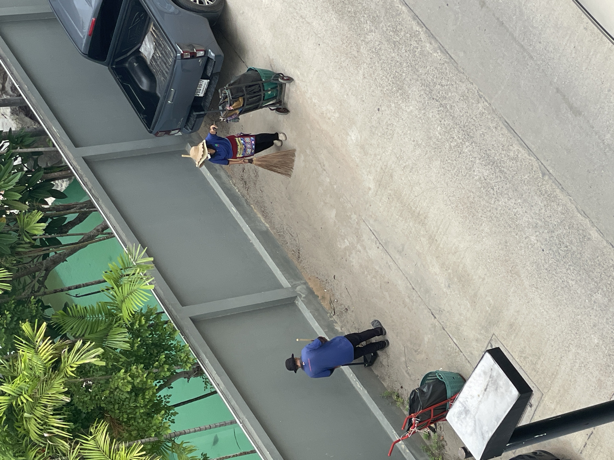 Two individuals are cleaning an outdoor paved area next to a grey concrete wall. The person on the left, wearing a blue shirt and a black hat, is holding a broom and dustpan, with a red wheeled cart containing a black trash bag and a green basket to their left. The person on the right, wearing a blue shirt, a straw hat, and a patterned apron, is sweeping with a broom and pushing a rolling cart with trash bags and other items. Behind them, a dark grey pickup truck is parked, with its license plate reading "2DD 8917". Lush green foliage and trees are visible behind the wall. This scene is located in Pattaya City, Thailand.