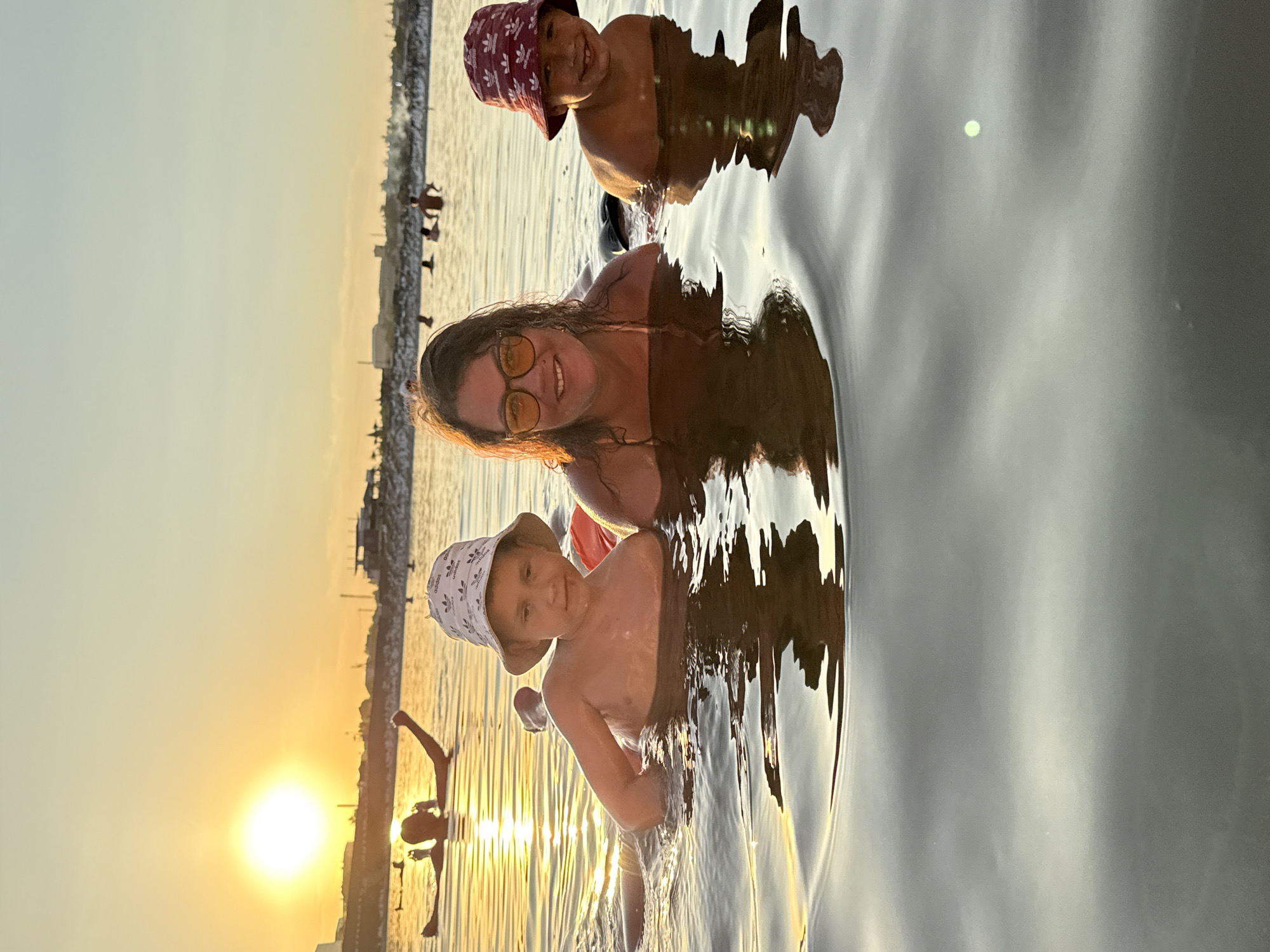 In the city of Sol-Iletsk, Russia, a group of people are seen swimming in a body of water during sunset. The sun, a bright yellow orb, is low on the horizon, casting a warm glow across the scene and reflecting off the rippling water. In the foreground, a woman wearing sunglasses and a pink bathing suit is visible, smiling at the camera. To her left, a young boy wearing a white bucket hat with an Adidas logo is also in the water, looking towards the viewer. To the right of the woman, another young boy is partially visible, wearing a red bucket hat. In the background, several other individuals are swimming and enjoying the water. The shoreline is lined with structures and vegetation. The overall atmosphere is one of leisure and enjoyment.