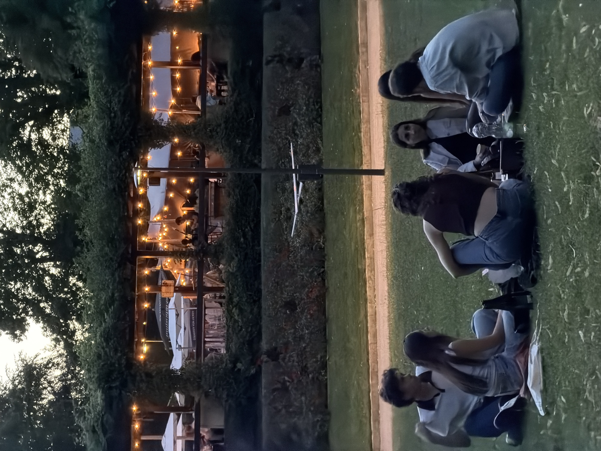 In the city of Luxembourg, Luxembourg, an outdoor dining area with string lights is visible in the background. In the foreground, six people are sitting on the grass, facing away from the camera. The group appears to be having a picnic or enjoying a casual gathering. The lighting suggests it is evening.