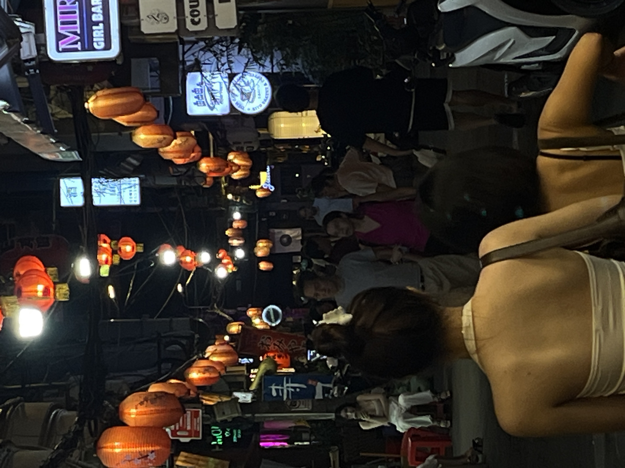 The image depicts a bustling street scene at night, likely in the city of Thủ Đức, Vietnam, characterized by its vibrant atmosphere and abundant red lanterns illuminating the narrow pathway. Several individuals are present, some walking, others standing. In the foreground, the backs of two women are visible, one wearing a white halter top and the other a black top. To the right, a man in a black t-shirt and shorts walks away from the camera, while a woman in a pink top stands to his left. A man in a light blue t-shirt is further back, to the left of the women in the foreground. The street is lined with various signs, some with neon lights, suggesting a commercial area with shops and establishments. Motorcycles are also visible, indicating common modes of transportation in the region. The overall impression is that of a lively night market or entertainment district.