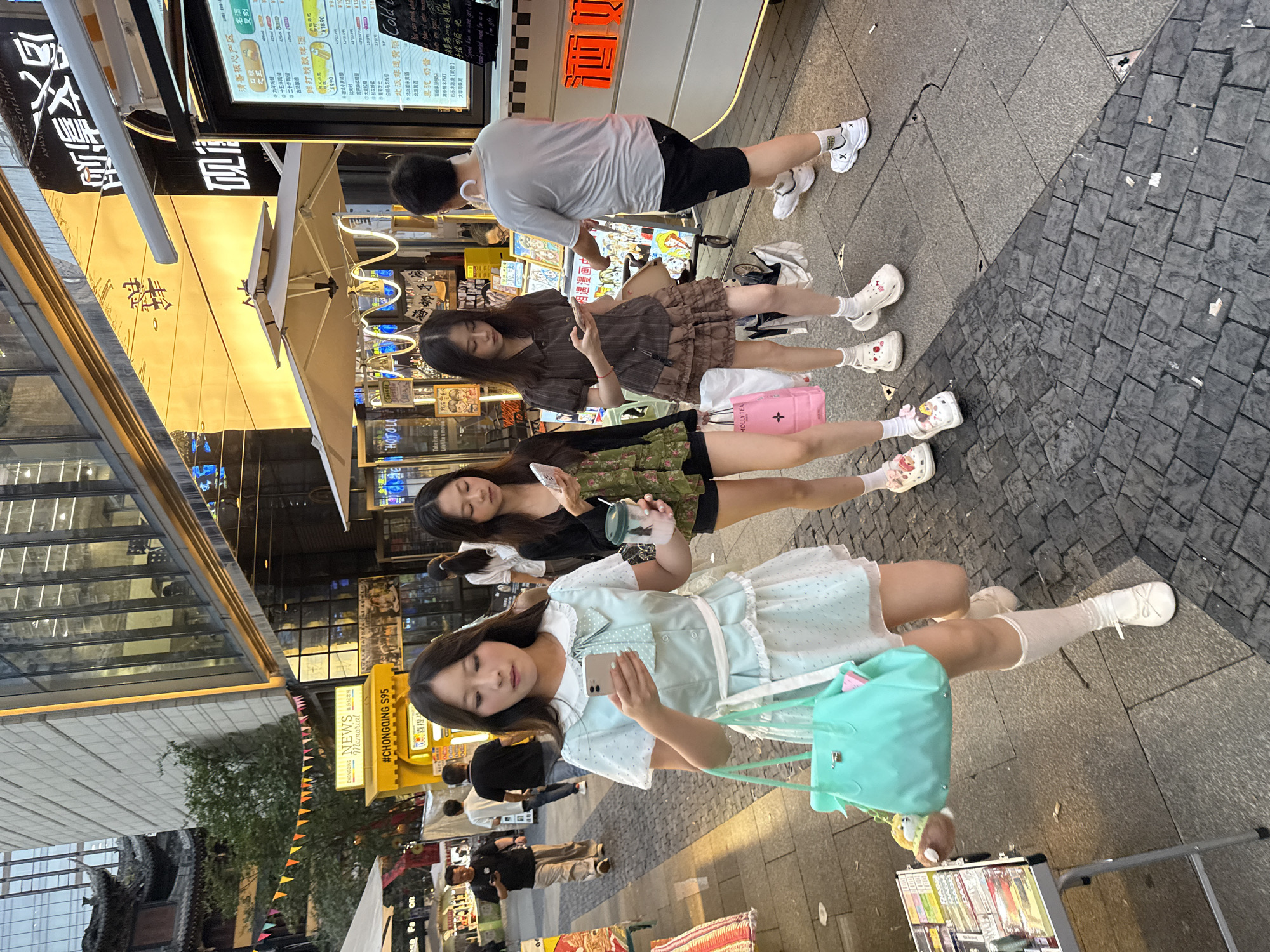 In the Yuzhong District of China, three young women are walking on a sidewalk lined with shops. The woman in the foreground, wearing a light blue polka dot dress and carrying a teal handbag, is looking at her phone. To her left and right, two other women are also looking at their phones, one holding a drink. In the background, a man in a gray t-shirt and black shorts walks away from the camera, while other pedestrians are visible further down the street. The scene is brightly lit by the setting sun, casting long shadows on the textured pavement. A yellow booth with "NEWS CHONGQING S95" written on it stands to the left of the women.