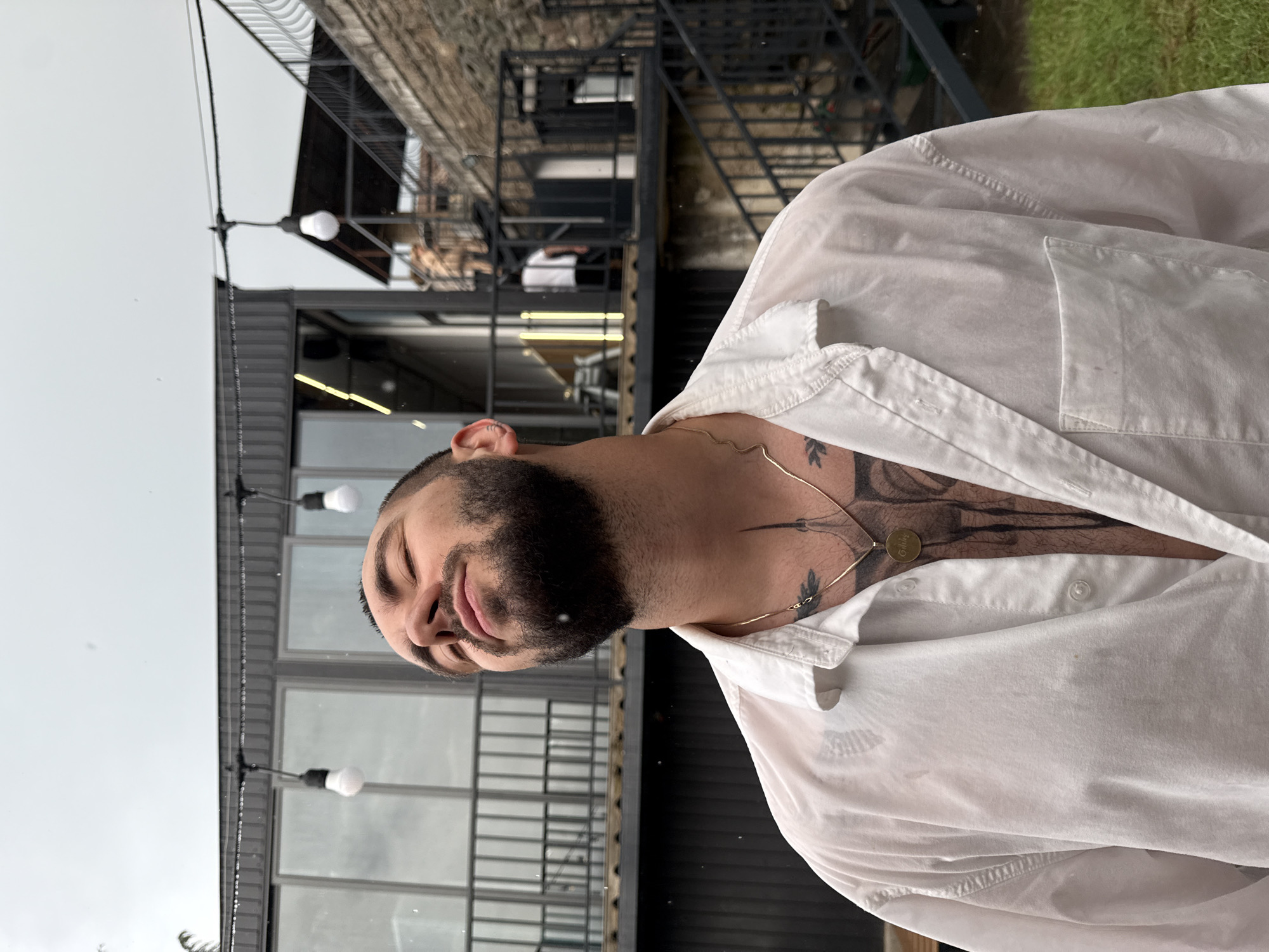 A man stands outdoors with his eyes closed and head tilted back, appearing to feel the rain. He wears an unbuttoned, wet, white shirt that reveals tattoos on his chest and neck. A gold chain with a pendant hangs around his neck. The background shows a modern building with large windows, a metal staircase, and some brickwork, consistent with the architectural style found in Dilijan, Armenia. String lights with exposed bulbs hang overhead, and raindrops are visible in the air.