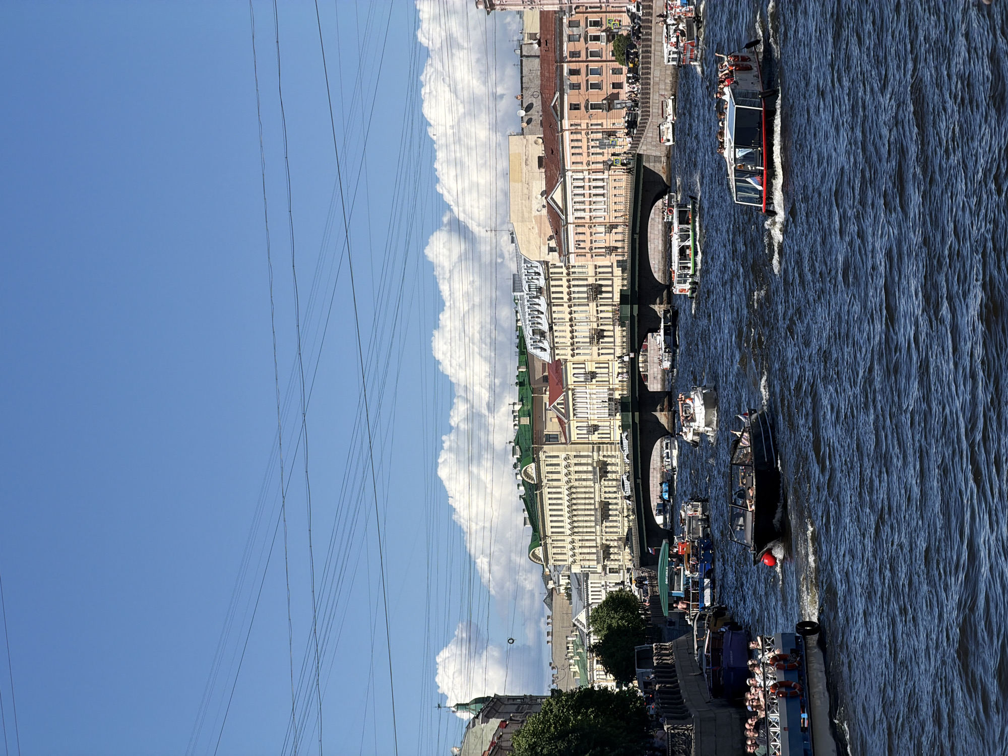 The image depicts a bustling waterway scene in Saint Petersburg, Russia, under a clear blue sky dotted with scattered white cumulus clouds. Numerous dark, thin power lines or communication cables crisscross the upper portion of the sky.
The foreground is dominated by dark blue, rippling water with visible white wakes from moving vessels. Multiple boats navigate the waterway. In the mid-ground, a dark-hulled motorboat with a red spherical buoy on its bow moves from right to left, creating a prominent wake. Towards the right foreground, a semi-enclosed tour boat, predominantly white with red accents and displaying a Russian flag, carries approximately ten passengers, some of whom are standing. On the left side, the stern of a larger, blue and white boat with numerous passengers visible on its deck is partially cropped. Several other tour boats, varying in size and design, are scattered across the waterway, some in motion, others appearing to be docked or slowly maneuvering near the embankments. One boat near the left embankment has a green canopy and visible passengers.
The middle ground features a dark-colored, arched bridge spanning the waterway. Vehicles and pedestrians are visible on the bridge deck. Both sides of the waterway are lined with stone embankments, along which people are walking. The left embankment features a row of trees.
In the background, a dense urban cityscape unfolds with multi-story historic buildings exhibiting classical architectural styles. Many buildings are light yellow or cream-colored, some with green roofs, while others display hues of peach, pink, or muted grey. A prominent building on the far left has a distinct green dome or spire. The architecture is consistent with the historic center of Saint Petersburg, suggesting a location along one of its central rivers or canals, such as the Fontanka or Moika River.