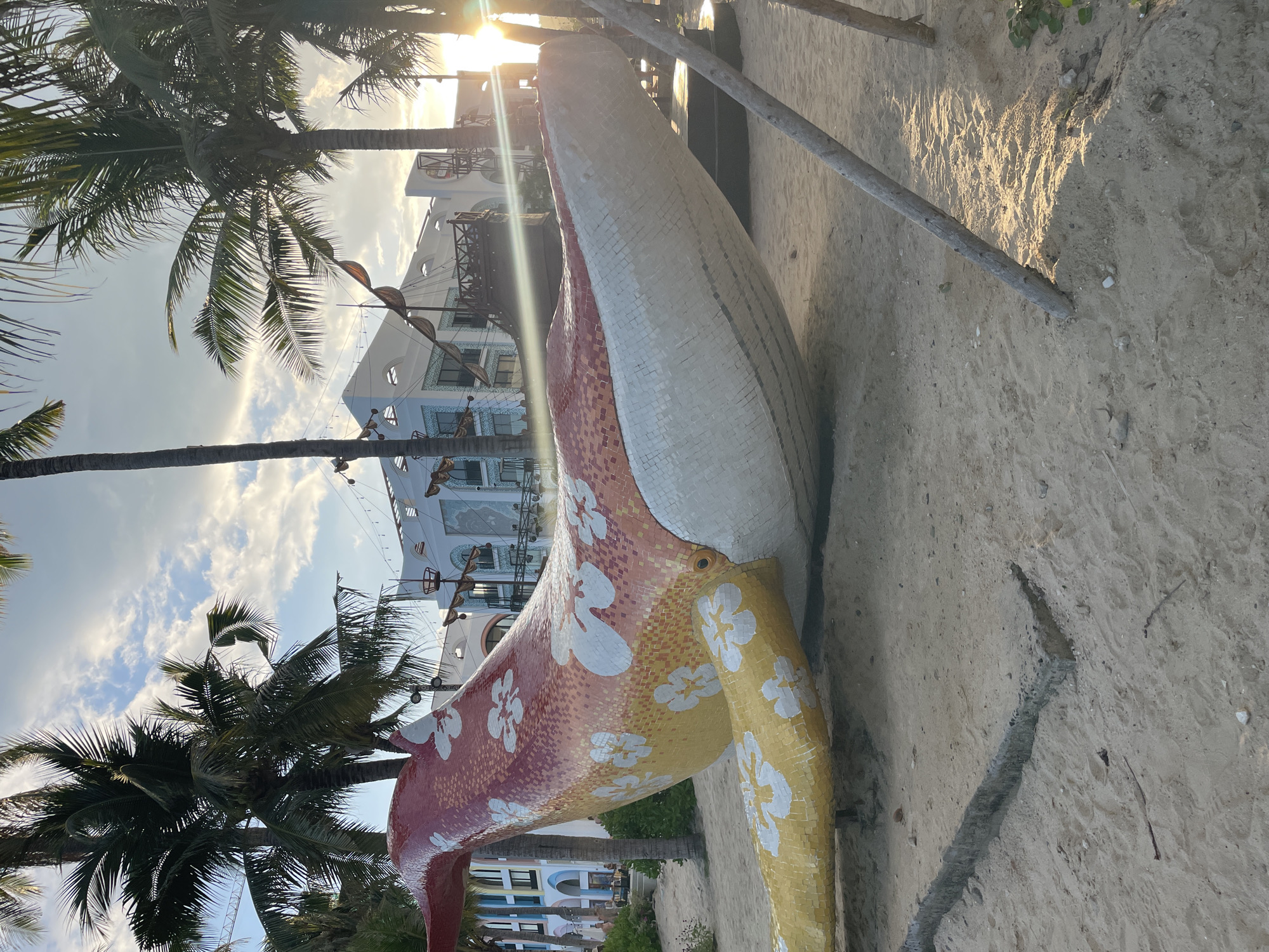 The image shows a large, mosaic-covered whale sculpture on a sandy beach. The whale is white on top and red/yellow on its body, with white floral designs. Palm trees are visible in the background, as well as a building with multiple balconies. Sunlight is visible, partially obscured by the whale's head. The setting appears to be in Bac Nha Trang Ward, Vietnam, based on the known location. The sand is light in color and appears dry. A wooden support is present next to the whale.