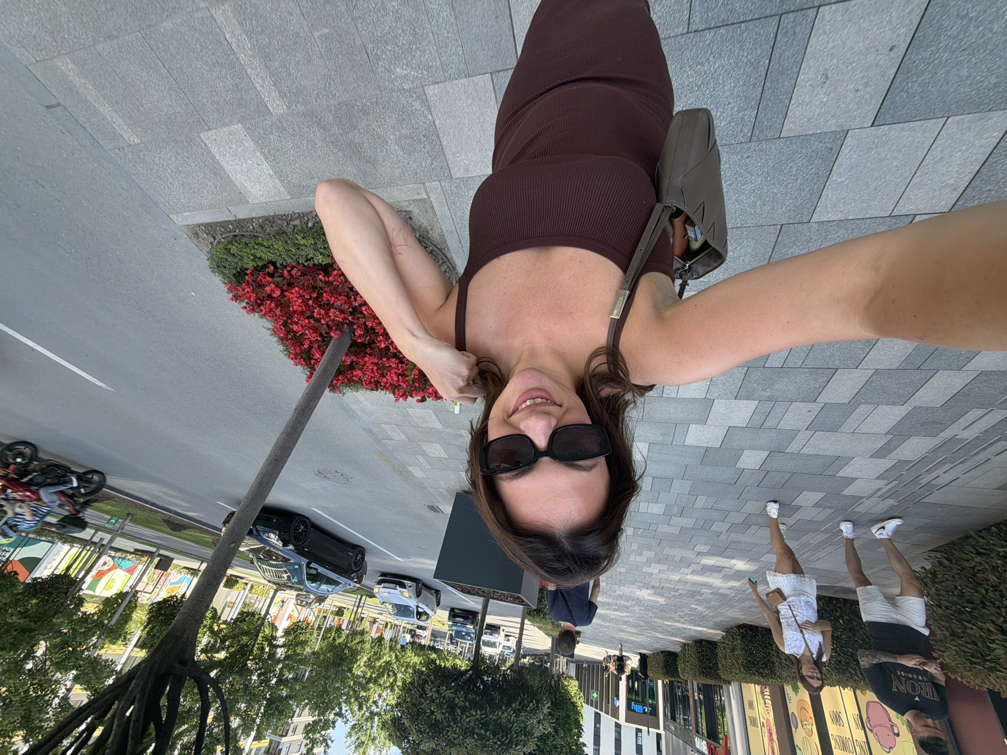 A wide-angle selfie shows a woman in a dark brown ribbed midi dress and dark sunglasses smiling as she walks on a sidewalk next to a street with traffic. Her brown shoulder bag hangs on her left shoulder. She is in an urban environment with modern buildings, trees lining the street, and planters with red flowers. In the background, other pedestrians are walking, and cars are driving on the street. The woman appears to be in Belgrade, Serbia, as indicated by the architectural style and the presence of a specific building with the "Lilly Drogerie" sign visible.