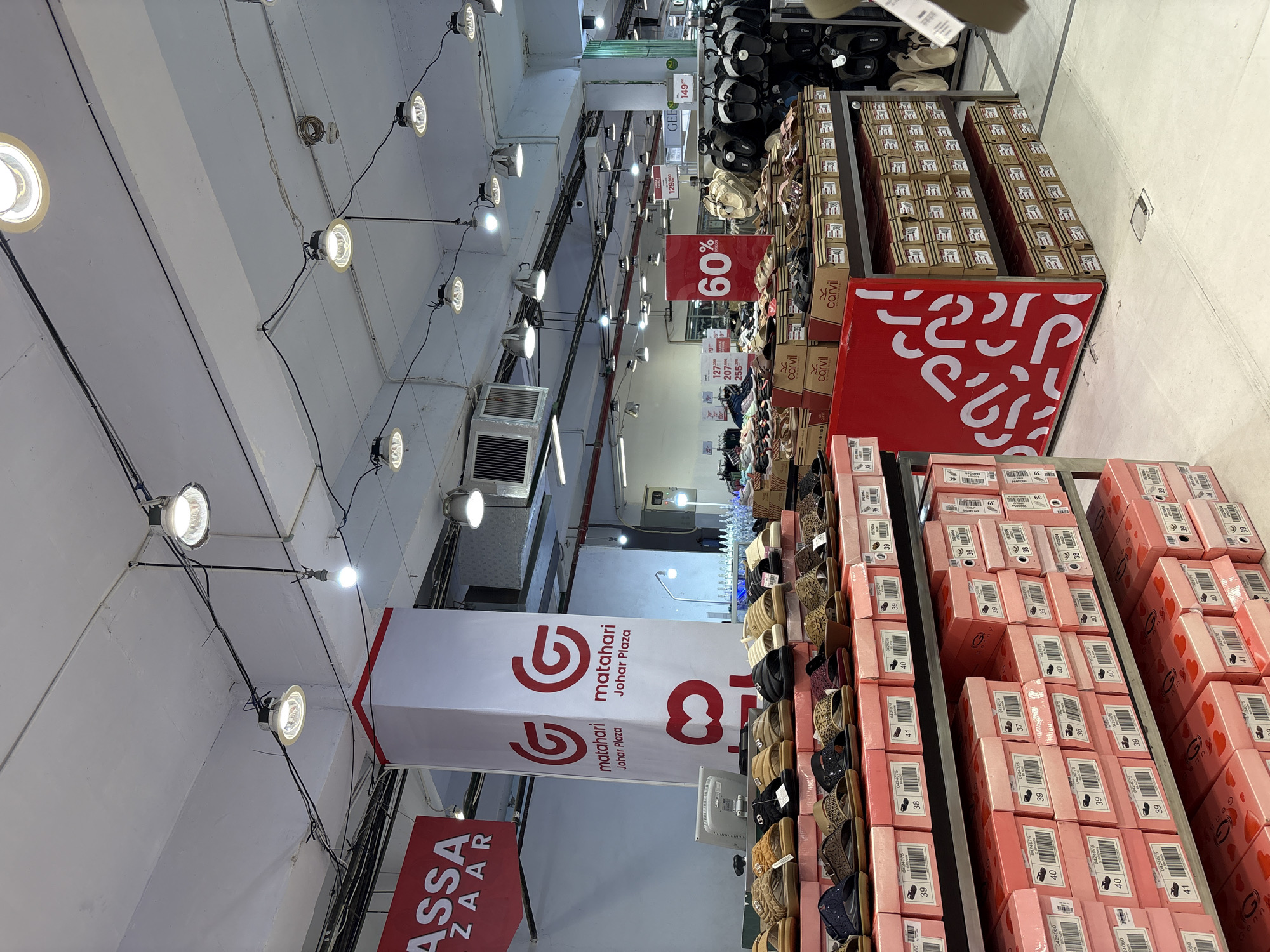 The image displays an indoor retail environment, likely a department store's shoe section, heavily stocked with merchandise and promotional signage. The view is angled upwards, showing the store's ceiling with exposed features and extensive shoe displays.
The ceiling is a functional white structure with visible concrete beams, numerous circular spotlights emitting bright light, fluorescent strip lights, and exposed electrical conduits. Several white air conditioning units are mounted high on the walls.
In the mid-ground, large vertical banners are prominent. A white banner with red text and a logo repeatedly reads "matahari Johar Plaza," indicating the store and its specific branch. Another red banner, partially visible in the lower left, displays "ASSA ZAAR."
The right side of the image is dominated by various shoe displays. Numerous pairs of sandals, slippers, and possibly heels, in colors such as black, beige, brown, and metallic, are neatly arranged on multi-tiered shelves. Adjacent to these are stacks of shoe boxes. Many boxes are pink with heart patterns and the brand "Genn" printed on them, while others are brown or beige with the brand "Carvil." Labels on the boxes clearly show shoe sizes including "37," "38," "39," "40," and "41," along with product codes like "0424075."
Promotional signs are also visible. A large red square sign with "60%" in white lettering indicates a discount. Smaller price tags display figures such as "129.800" and "149," which are likely prices in Indonesian Rupiah, consistent with the Jember, Indonesia location. One tag also shows the text "GEE." A red display barrier on the lower right features a repeating white, abstract pattern resembling inverted question marks.
The scene conveys a busy, sale-oriented atmosphere within a large retail outlet in Jember, Indonesia.
