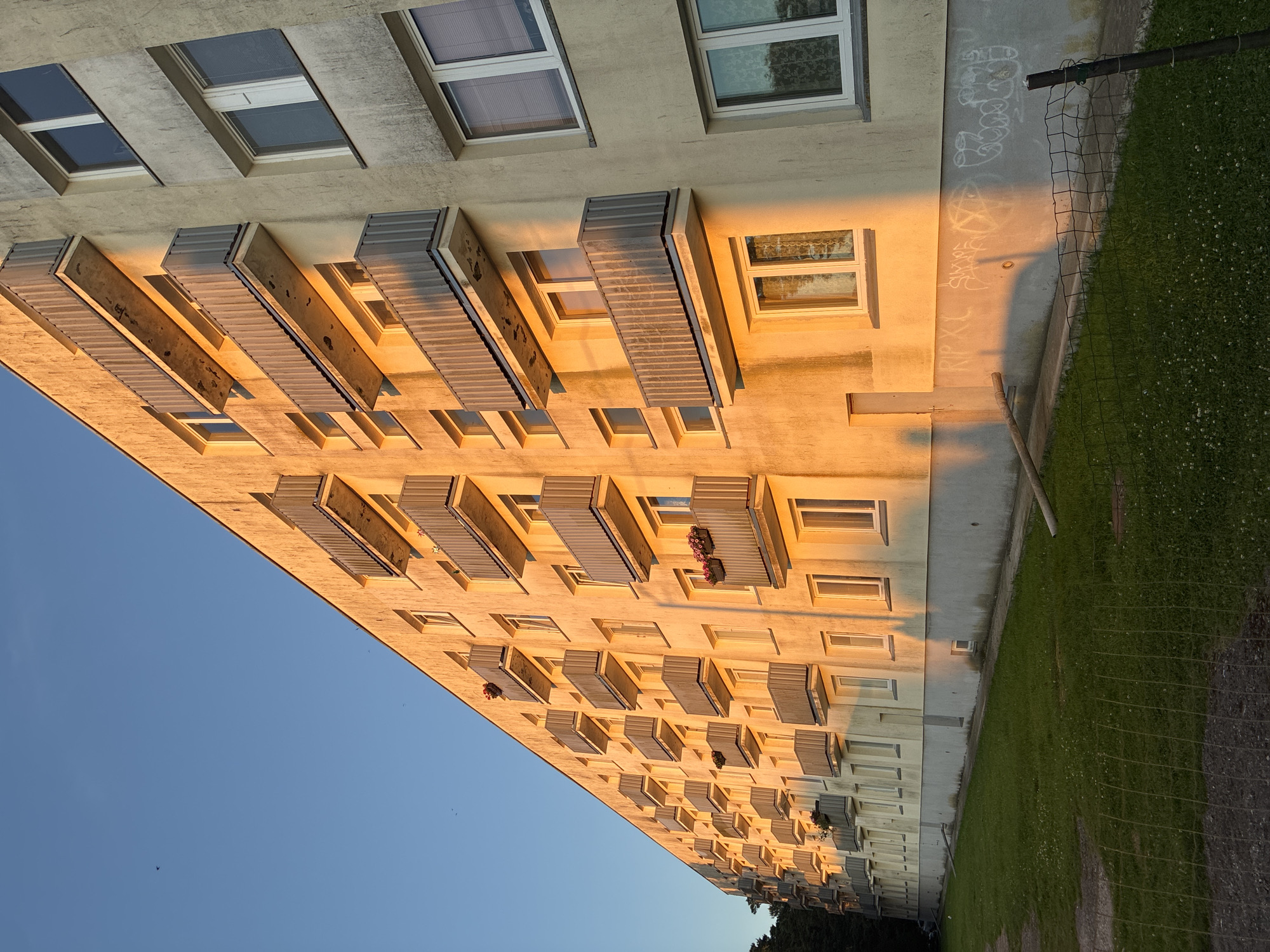 The image shows a multi-story apartment building in Haapsalu, Estonia, bathed in the warm golden light of the setting sun. The building features rows of balconies with metal railings and concrete slabs. The facade is painted a light yellow and shows signs of age and some staining. Several windows with blinds are visible. The ground level of the building has a strip of concrete with visible graffiti. In front of the building, there is a well-maintained grassy area with some scattered white flowers. A wire fence with a metal post runs along the foreground, separating the lawn from what appears to be a dirt path. The sky above is a clear, bright blue. There are no people visible in the image.