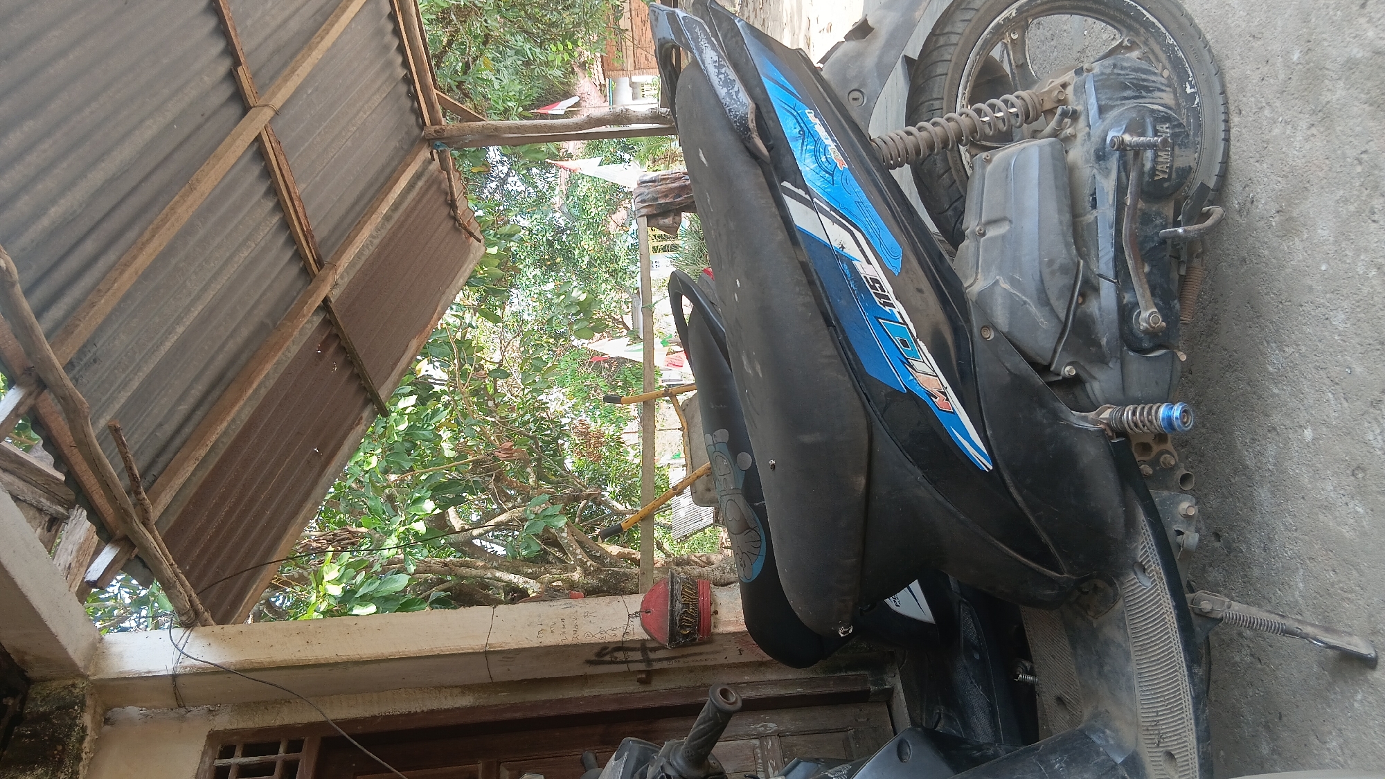 In the city of Prabumulih, Indonesia, under a weathered corrugated metal roof supported by wooden beams, two black scooters are parked on a concrete surface. The scooter in the foreground displays blue and white decals with the text "MIO 115". Its engine is visible, with the "YAMAHA" logo clearly imprinted on the casing. A silver kickstand is extended on the left side of the scooter. In the background, trees with green foliage are visible, and two Indonesian flags are fluttering in the distance. The scene appears to be an outdoor, shaded area, possibly a carport or a similar shelter.