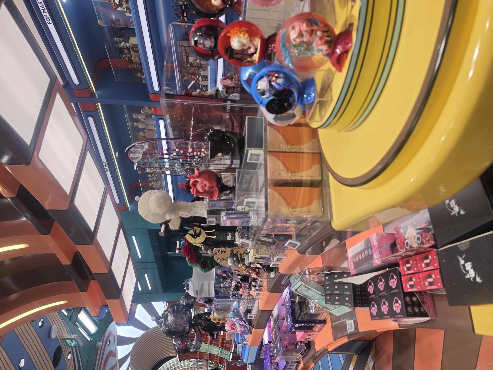 The image depicts the interior of a Pop Mart store, likely in Bangkok, Thailand, given the store's presence in the city. The image is taken from a low angle, tilted to the left, showcasing the store's layout with shelves and displays filled with collectible toys. The ceiling is decorated with colorful horizontal stripes and lighting fixtures.
On the right, a yellow display unit with colorful collectible figures in plastic bubbles is prominent. Shelves filled with boxed collectible toys are visible below this unit. Further back, more shelves display various toys and figures, including larger plush characters. The store seems well-lit and filled with merchandise, typical of a retail environment. No people are visible in the frame.