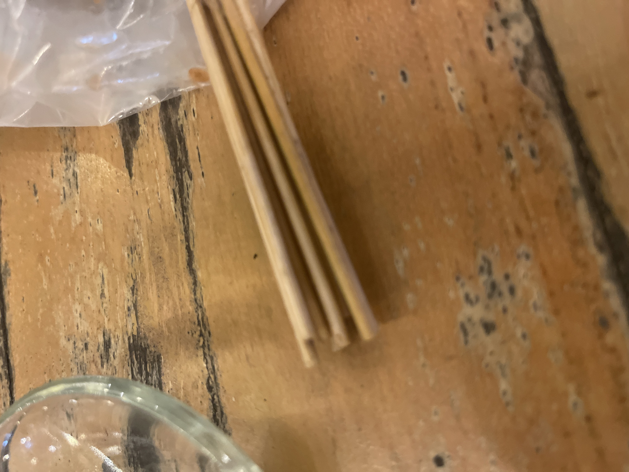 The media file is a close-up, top-down shot of wooden chopsticks in a plastic wrapper, lying on a textured wooden surface. The chopsticks are a light brown or beige color and appear to be made of bamboo or a similar wood. They are bundled together, with the wrapper covering the top portion of them. The wooden surface has a distressed look with visible grain and dark markings, possibly stains or natural imperfections.
In the lower left corner of the image, a portion of a clear glass, possibly a bowl or cup, is visible. The glass has a curved rim and contains some small bubbles. The lighting in the image is somewhat diffused, casting soft shadows.
The scene appears to be an indoor setting, given the focus on objects on a table. There are no people visible in the frame. The overall impression is one of a simple, perhaps domestic or restaurant setting, focusing on food-related items. The image does not suggest a specific time of day or weather conditions. There is no visible text in the media file.