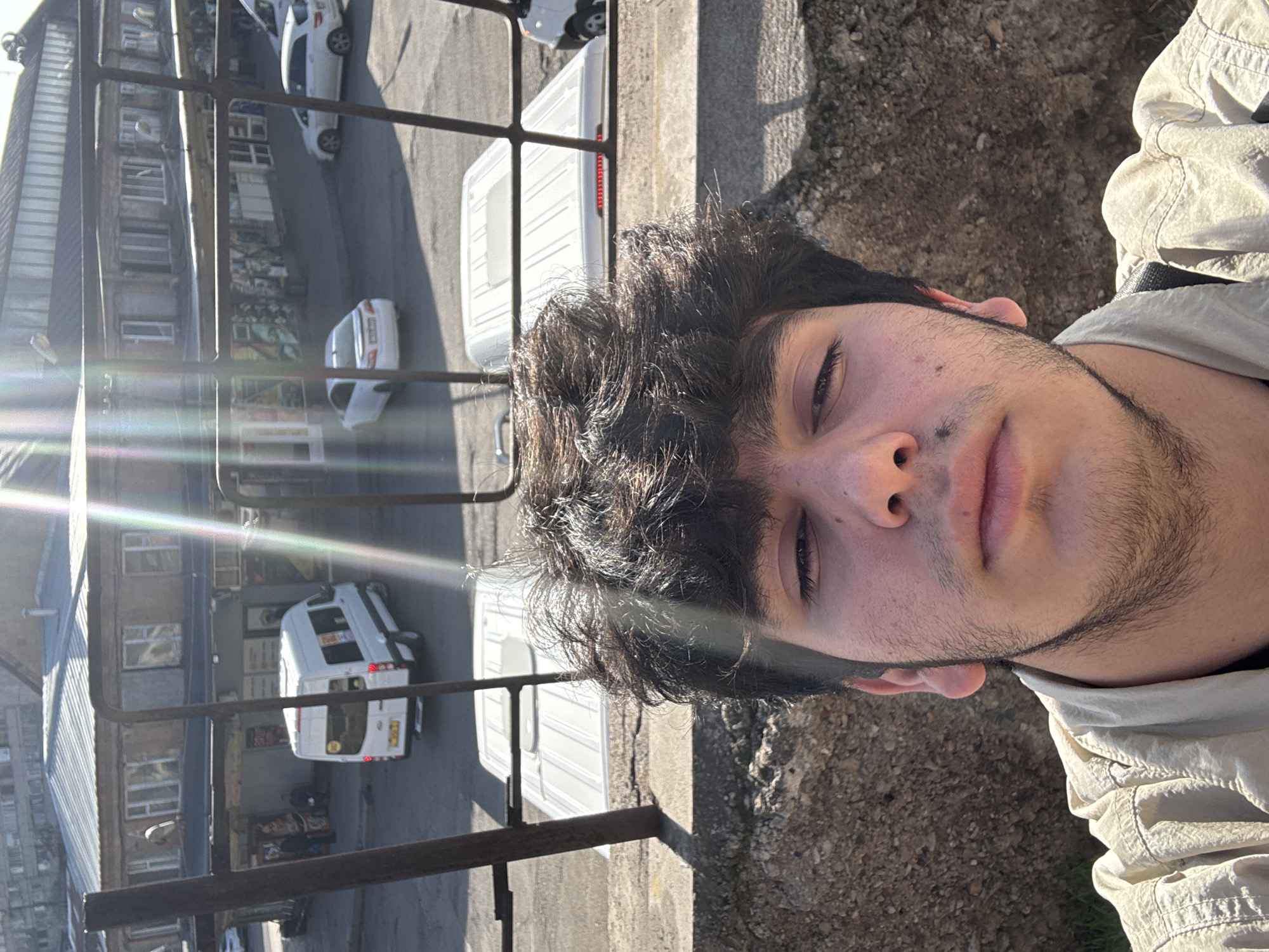 The image is a selfie taken by a young man with dark, curly hair and stubble. He is wearing a light-colored jacket or shirt. The background shows an urban scene, possibly a street or courtyard, observed from an elevated position. There are buildings with multiple floors, and several white vans or small trucks are parked on the street. The perspective is angled downwards, and there are metal railings in the foreground, suggesting a balcony or platform. The location appears to be Yerevan, Armenia, as the architectural style and the presence of vans are often seen in that city. There is strong sunlight, causing lens flare in the image. The man is looking into the camera with a neutral expression.