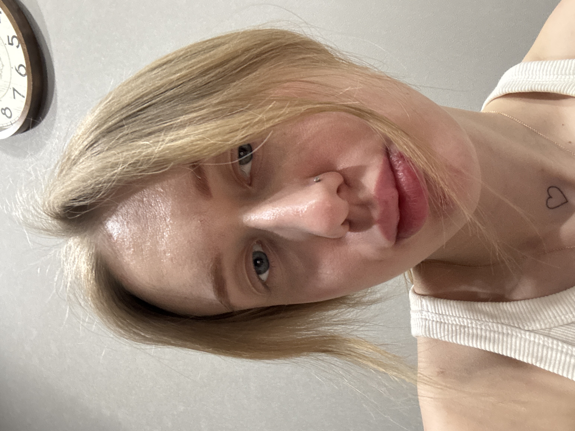 A close-up, head-and-shoulders portrait depicts an individual, likely female, with fair skin and light blonde hair that falls over the forehead and partially obscures the left side of the face. The subject has blue eyes and a small silver stud piercing on the right nostril. The lips appear full, with a natural sheen. The subject wears a white ribbed tank top, revealing a small outline heart tattoo on the upper chest, just below the collarbone, and a thin gold-colored chain necklace. The background is a plain, light grey wall. In the upper right corner, a portion of a round, wooden wall clock is visible, displaying the numbers 8, 7, 6, and 5. The specific location within Rostov-on-Don, Russia, cannot be determined from the visual information present in the image.
