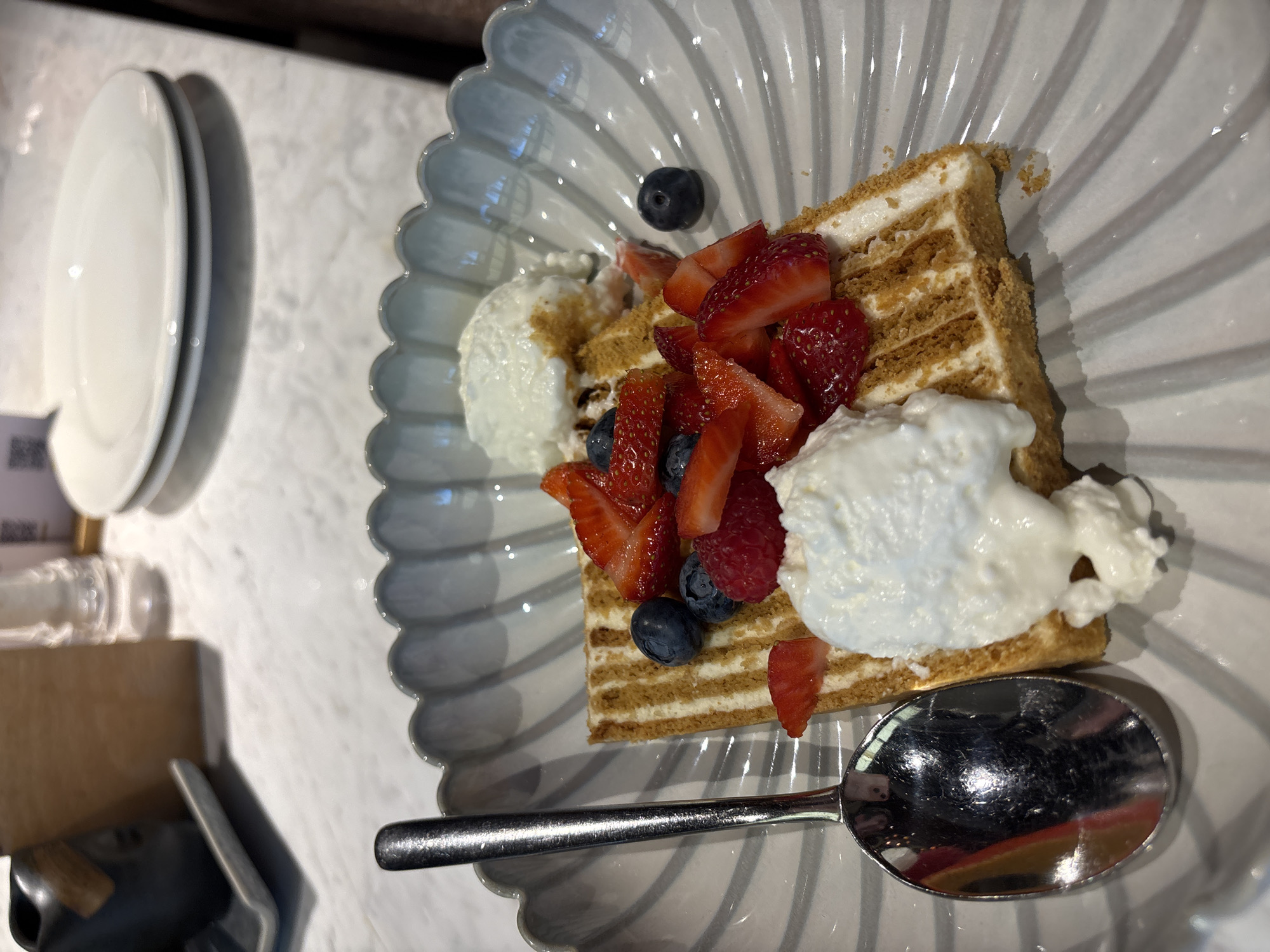 A dessert is presented in a fluted grey ceramic bowl. The dessert is a layered cake with visible cream and cookie crumbs. It is topped with fresh strawberries, raspberries, and blueberries. Two dollops of whipped cream are placed on top of the dessert. A silver spoon rests to the left of the dessert, positioned inside the bowl. The background shows a white marbled table, two white plates stacked on top of each other, a glass shaker, and a dark ceramic object with a wooden label. The scene is likely at a cafe or restaurant in Saint Petersburg, Russia, based on the overall aesthetic and the presence of typical dining ware.