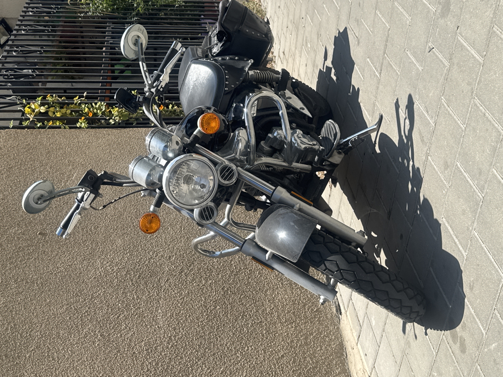The image shows a motorcycle parked on a brick sidewalk in Tel-Aviv, Israel. The motorcycle is a cruiser style, with a black studded seat, chrome accents, and a large headlight. The motorcycle is parked next to a wall on one side and a fence with greenery on the other. The sunlight casts a shadow of the motorcycle onto the sidewalk.