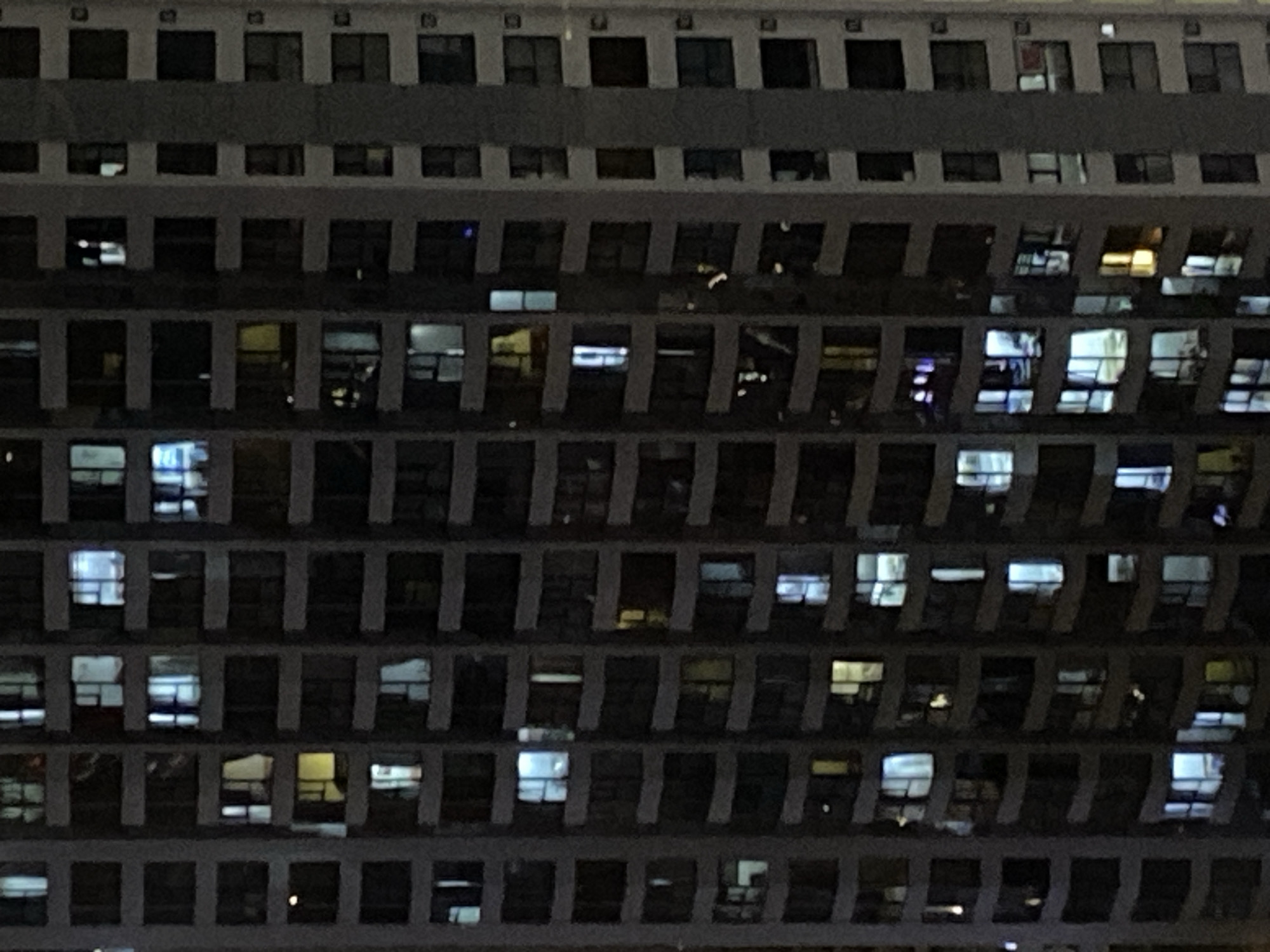 The image shows a close-up view of a residential building in Nha Trang Ward, Vietnam, at night. The building is tall and features multiple floors with numerous windows. Many of the windows are lit from within, indicating that people are inside and awake. The lighting varies from a warm yellow to a cooler blue, suggesting different activities or types of lighting within the apartments. The architectural style appears to be modern, with balconies visible on some floors. The overall impression is of a bustling urban environment, with the lit windows creating a pattern against the dark facade of the building. The specific location is an apartment building in Nha Trang Ward, Vietnam.