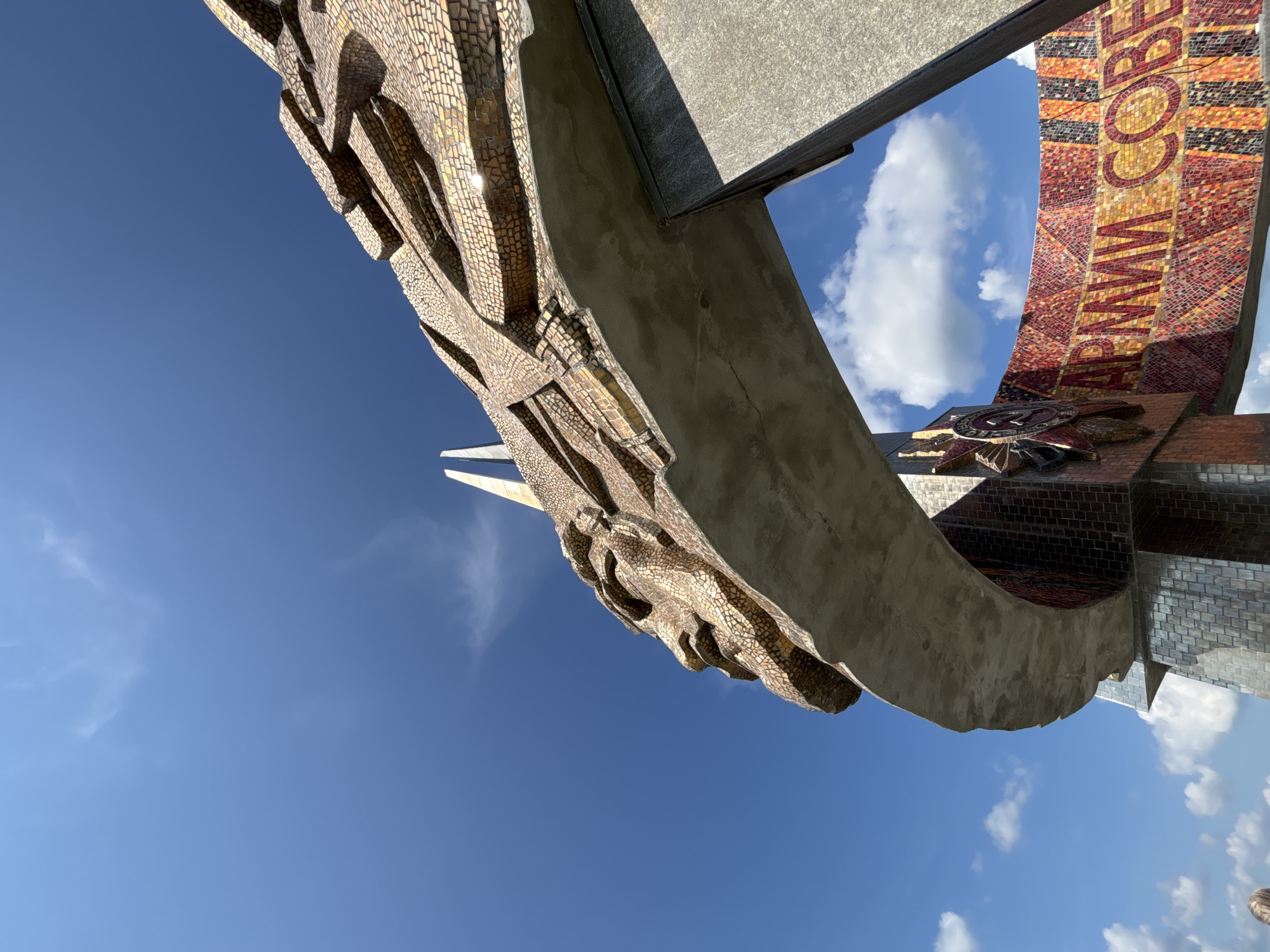 A low-angle, wide shot captures a monumental sculpture against a clear blue sky with sparse white clouds. The sculpture features a mosaic-covered face in profile, constructed from small, golden, irregular shapes. The face is part of a large, arched structure that extends across the frame. The arch is supported by granite pillars and decorated with a colorful mosaic band that wraps around its base. Red, orange, yellow, and black tiles form a repeating pattern, with some tiles appearing to be part of a Cyrillic inscription that reads "АРМИИ СОВЕ." A large, star-shaped emblem, likely a Soviet military award, is also visible on the structure. The sculpture appears to be a monument dedicated to the Soviet Army. The overall impression is one of grandeur and historical significance. The location is the Ozyaritska-Slabadski rural council in Belarus.